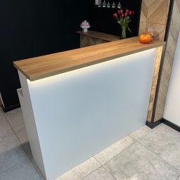 Mateusz Jakubczak - Nowoczesna recepcja z białym frontem i blatem w kolorze dębu pradawnego, podświetlona od spodu, z dekoracjami w postaci tulipanów, dyni i kwiatów, na tle czarnej ściany i ściany wykończonej...