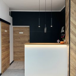 Mateusz Jakubczak - Recepcja z białym frontem podświetlonym ledami, drewnianym blatem, czarną ścianą w tle i trzema wiszącymi lampami. Po lewej stronie przesuwne drzwi z drewnianym wzorem, a po prawej ścianka...