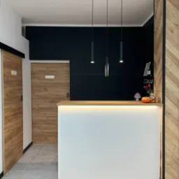 Recepcja z białym frontem podświetlonym ledami, drewnianym blatem, czarną ścianą w tle i trzema wiszącymi lampami. Po lewej stronie przesuwne drzwi z drewnianym wzorem, a po prawej ścianka...