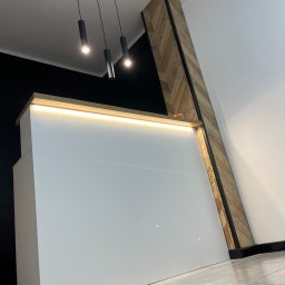 Mateusz Jakubczak - Biała, minimalistyczna zabudowa meblowa z podświetleniem LED i drewnianym akcentem w jodełkę, uzupełniona o czarne lampy sufitowe i szarą podłogę.