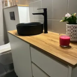 Czarna, okrągła umywalka nablatowa z czarną baterią na drewnianym blacie szafki łazienkowej z szufladami, obok świeczka i kwiat w ozdobnej doniczce.