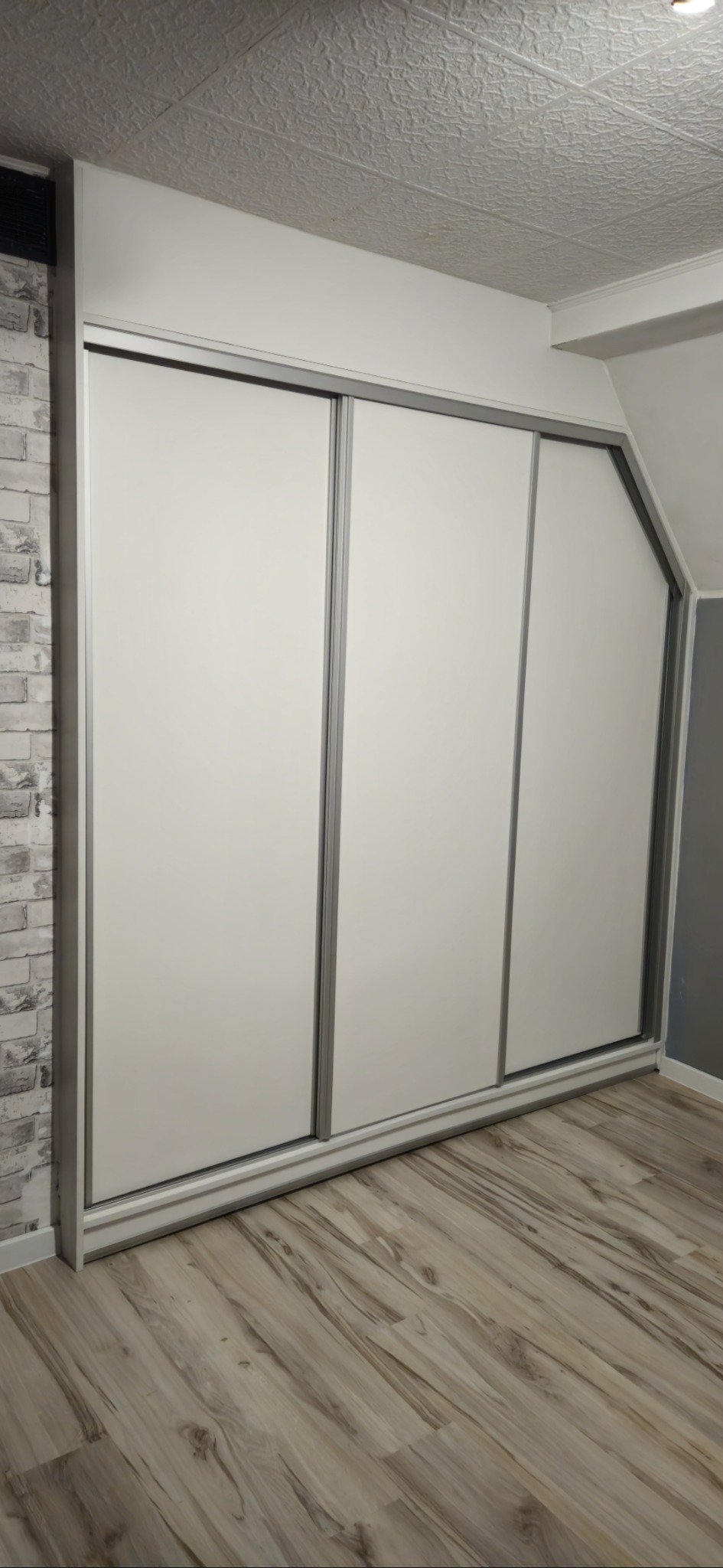 Trzydrzwiowa szafa przesuwna z białymi frontami i srebrnymi obramowaniami, wkomponowana we wnękę pod skosem sufitu, z widoczną ścianą z imitacji cegły.