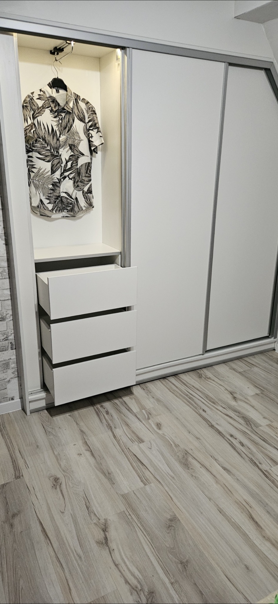 Biała szafa przesuwna z trzema wysuniętymi szufladami i koszulą w tropikalne wzory wiszącą w oświetlonej wnęce, na podłodze panele imitujące drewno.