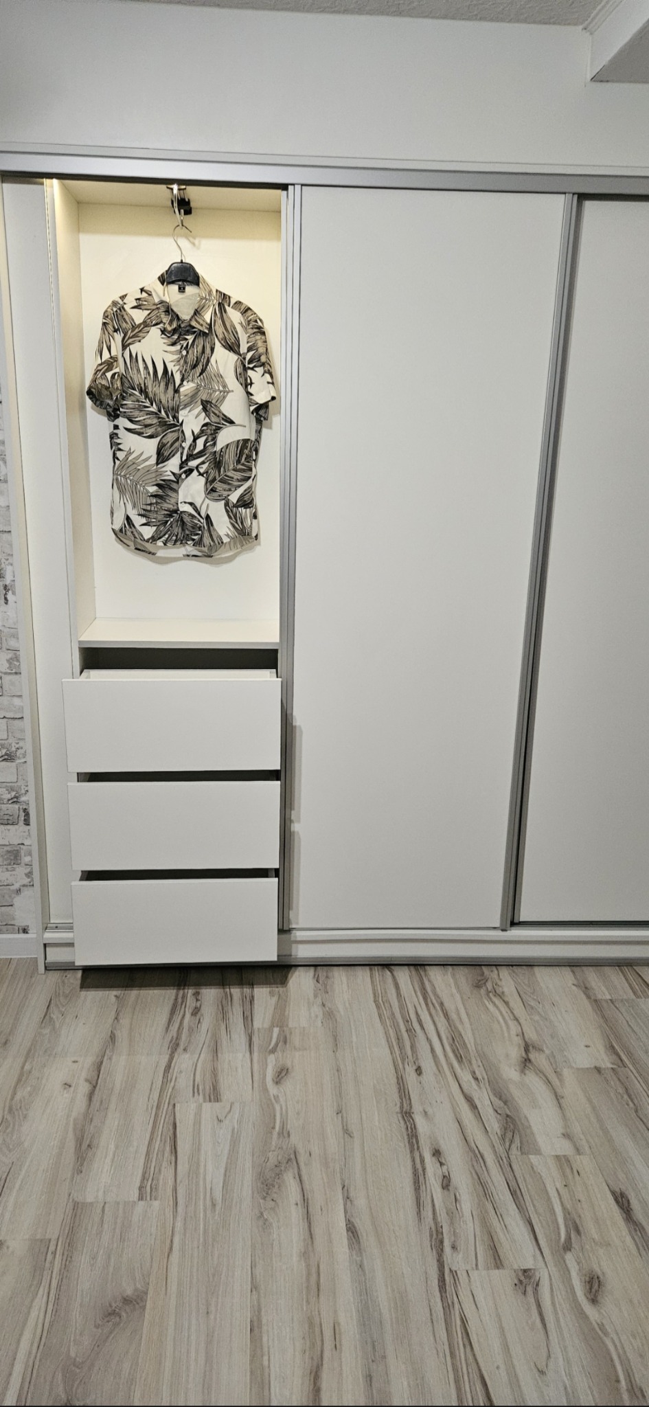 Biała szafa przesuwna z otwartą wnęką z trzema szufladami i koszulą w tropikalny wzór wiszącą na wieszaku, widoczna faktura drewnopodobnej podłogi.