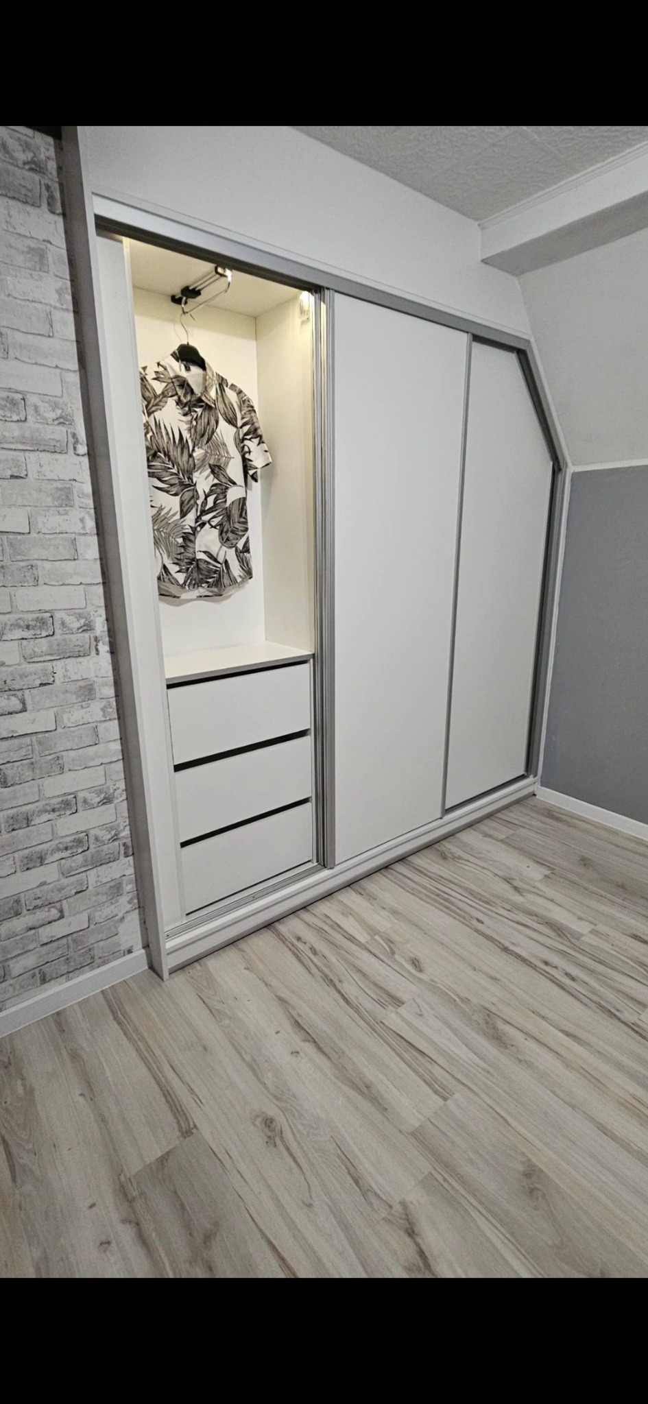 Biała szafa przesuwna z trzema drzwiami i szufladami, oświetlona od wewnątrz, z wiszącą koszulą w liście, na tle ściany z cegły i drewnianej podłogi.