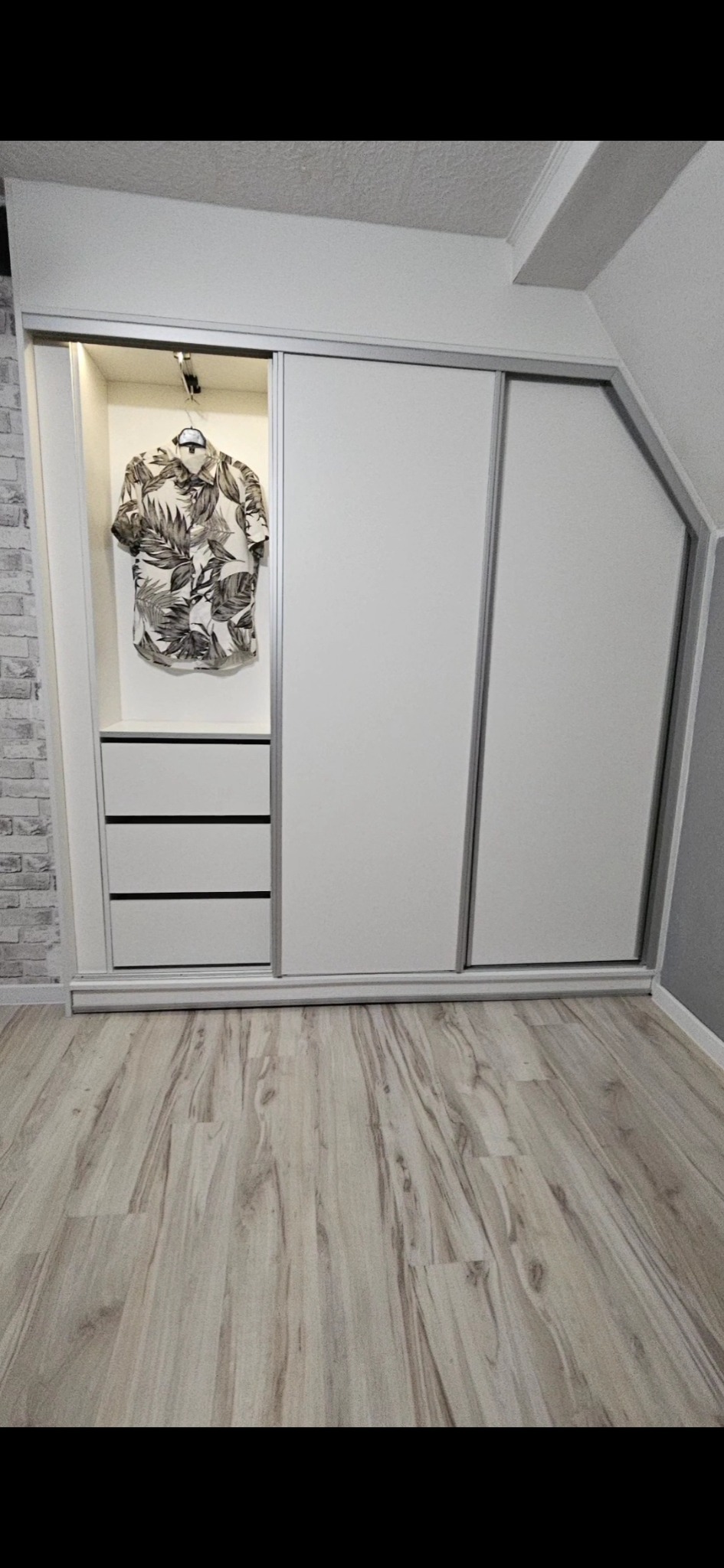 Biała szafa wnękowa z przesuwnymi drzwiami, trzema szufladami i koszulą w tropikalne liście wiszącą w otwartej części, na poddaszu z podłogą z paneli.