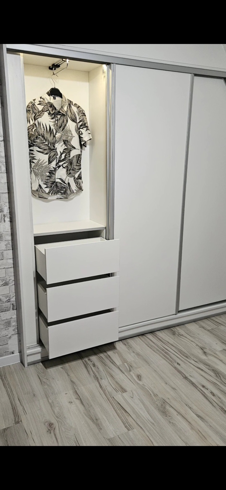 Biała szafa przesuwna z otwartym segmentem z szufladami i koszulą w tropikalne wzory na wieszaku, oświetlenie LED wewnątrz, panele podłogowe w odcieniach szarości.