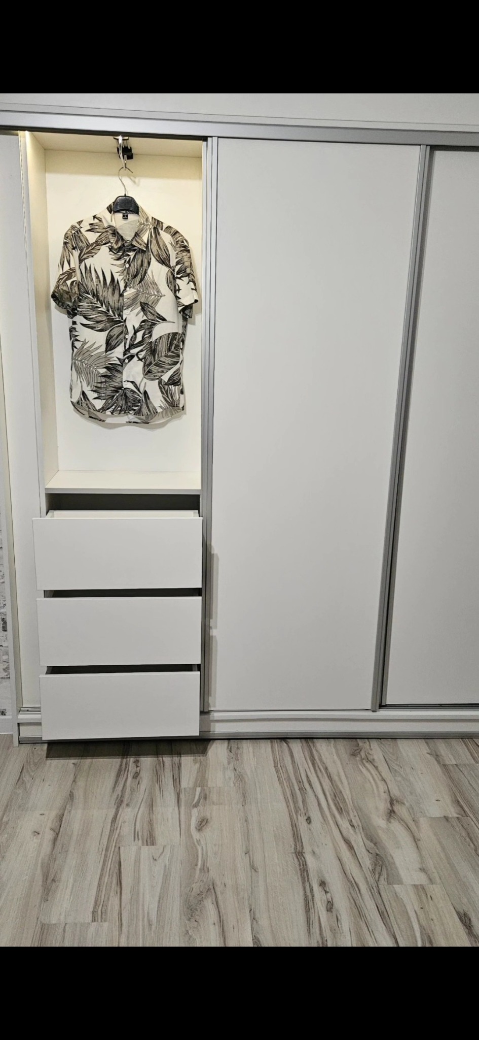 Biała szafa przesuwna z otwartą częścią z trzema szufladami i koszulą w tropikalne wzory na wieszaku.