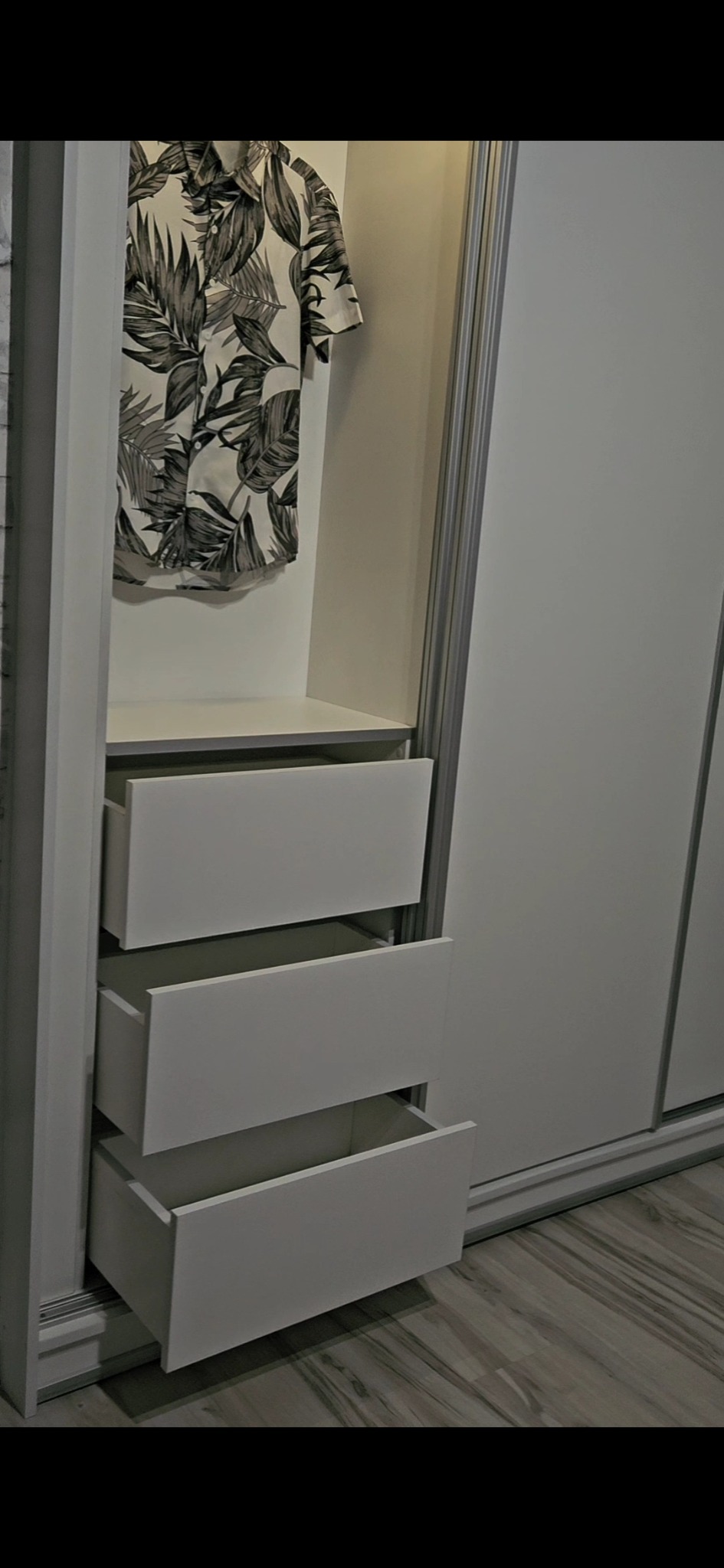 Biała szafa z przesuwnymi drzwiami i otwartymi szufladami, w górnej części widoczna koszula w liście.