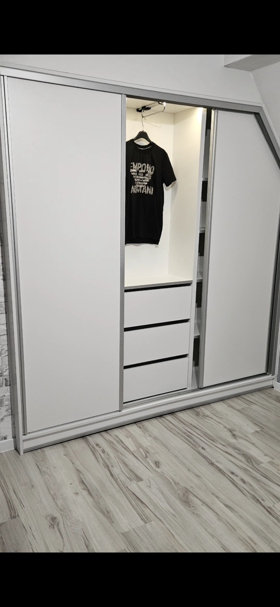 Biała szafa przesuwna z trzema szufladami, częściowo otwarta, z czarną koszulką na wieszaku w środku i jasną podłogą panelową.