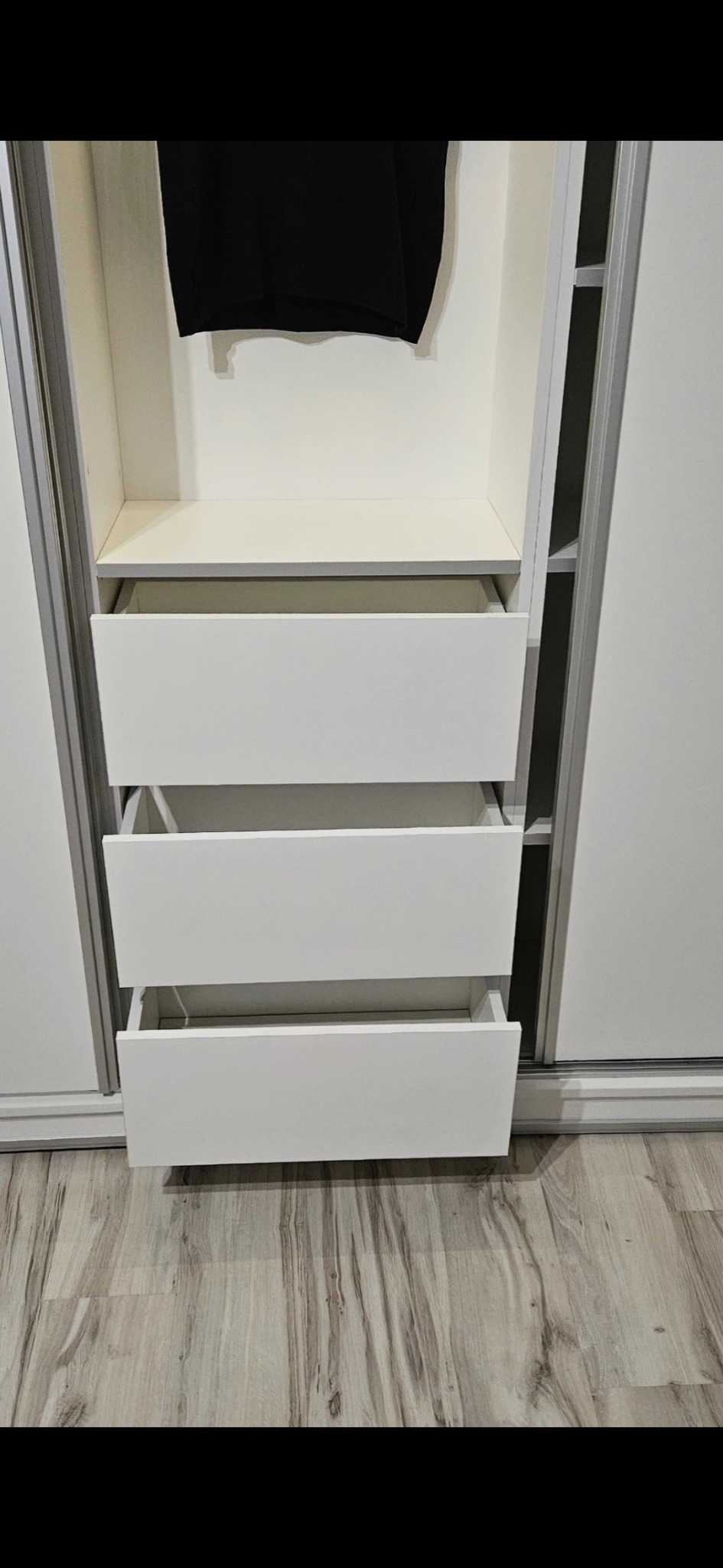 Wnętrze białej szafy wnękowej z trzema wysuwanymi szufladami, z widocznym fragmentem czarnego ubrania wiszącego na górze.