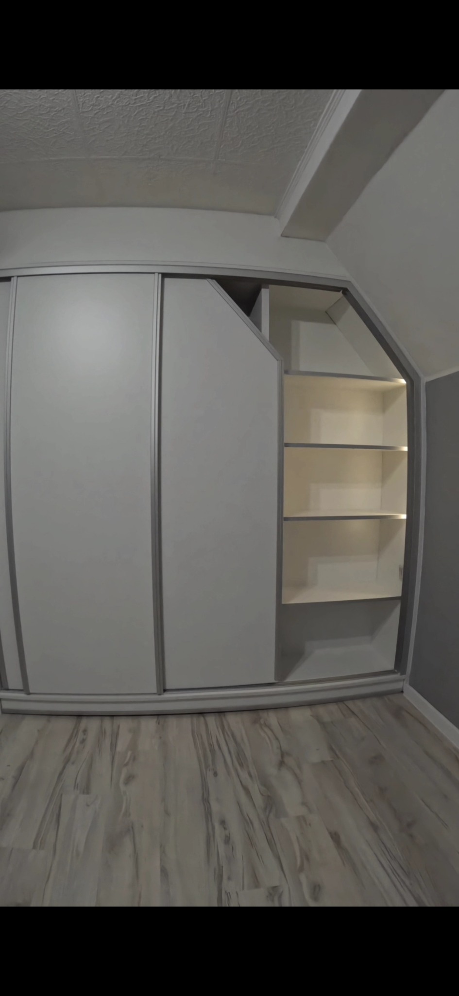 Biała szafa wnękowa z przesuwnymi drzwiami i otwartą częścią z półkami, dopasowana do skosu dachu, na jasnej podłodze imitującej drewno.
