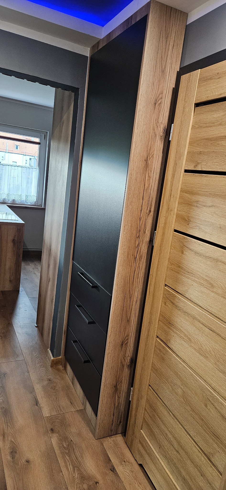 Wnętrze z zabudową: szafa z czarnymi frontami i trzema szufladami, obudowana drewnopodobną płytą, widoczna podłoga z paneli i fragment drzwi z drewnianą okleiną.