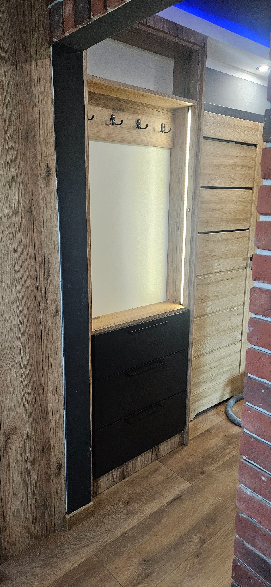 Wnęka z szafką na buty i wieszakami, wykończona w drewnie z trzema czarnymi szufladami i oświetleniem LED, widok z boku na korytarz z drewnianą podłogą i ceglaną ścianą.