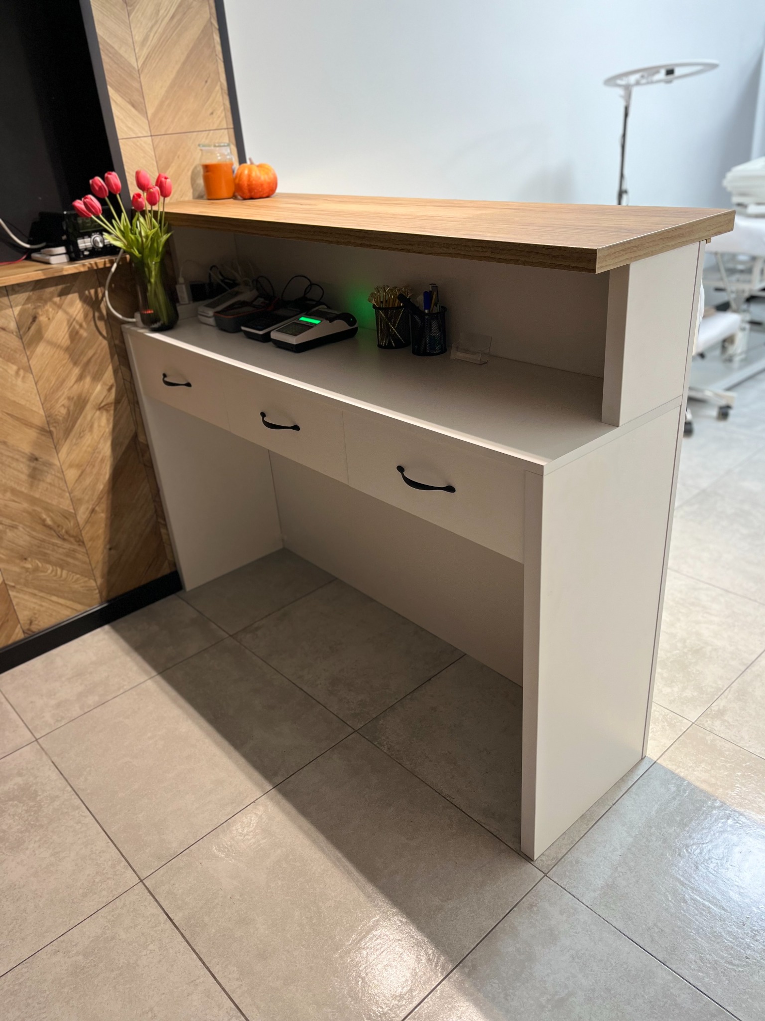 Biała lada recepcyjna z drewnianym blatem w salonie w Wałbrzychu, na blacie wazon z tulipanami i dekoracje, w tle sprzęt kosmetyczny.