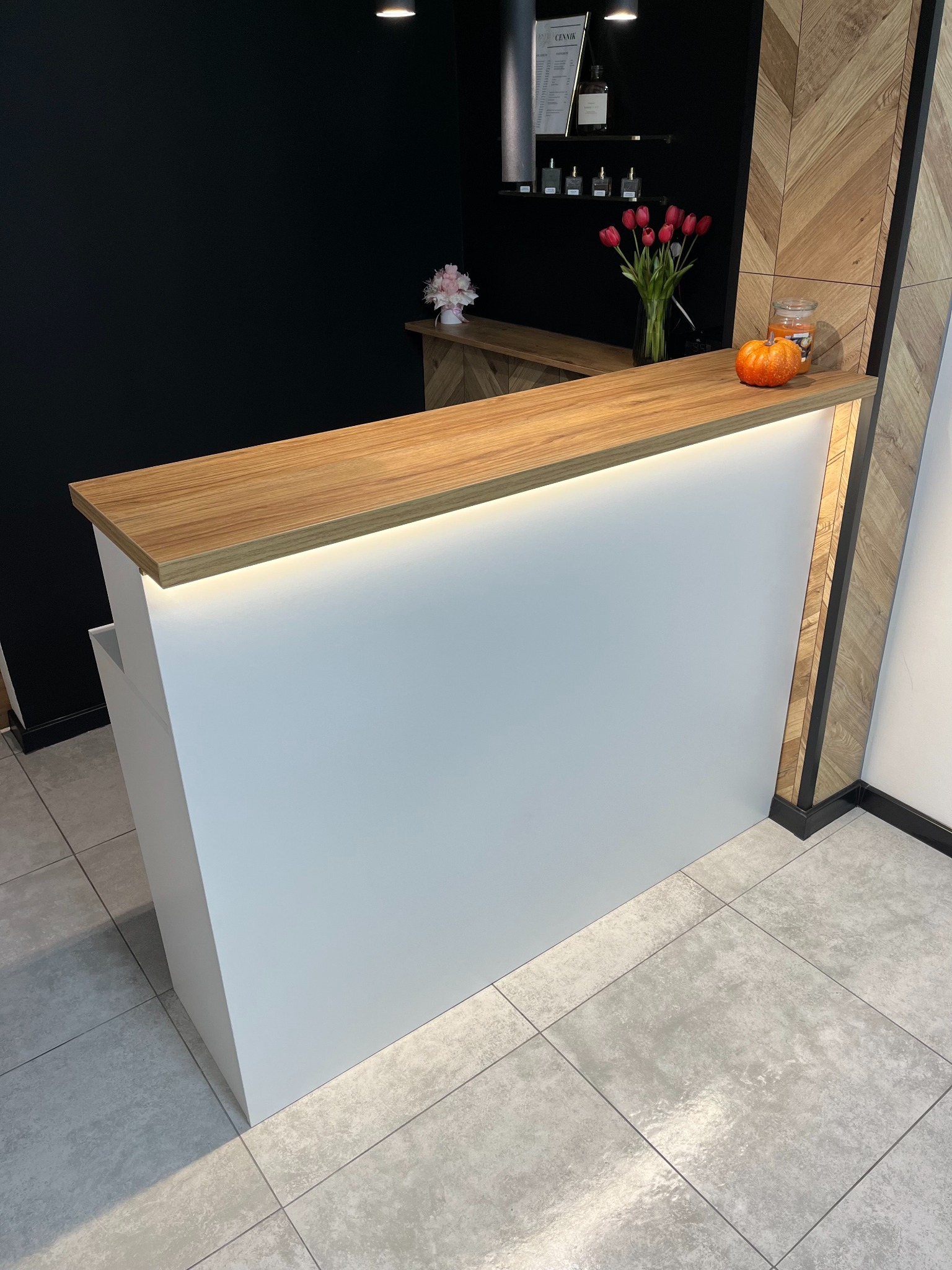 Nowoczesna recepcja z białym frontem i blatem w kolorze dębu pradawnego, podświetlona od spodu, z dekoracjami w postaci tulipanów, dyni i kwiatów, na tle czarnej ściany i ściany wykończonej...
