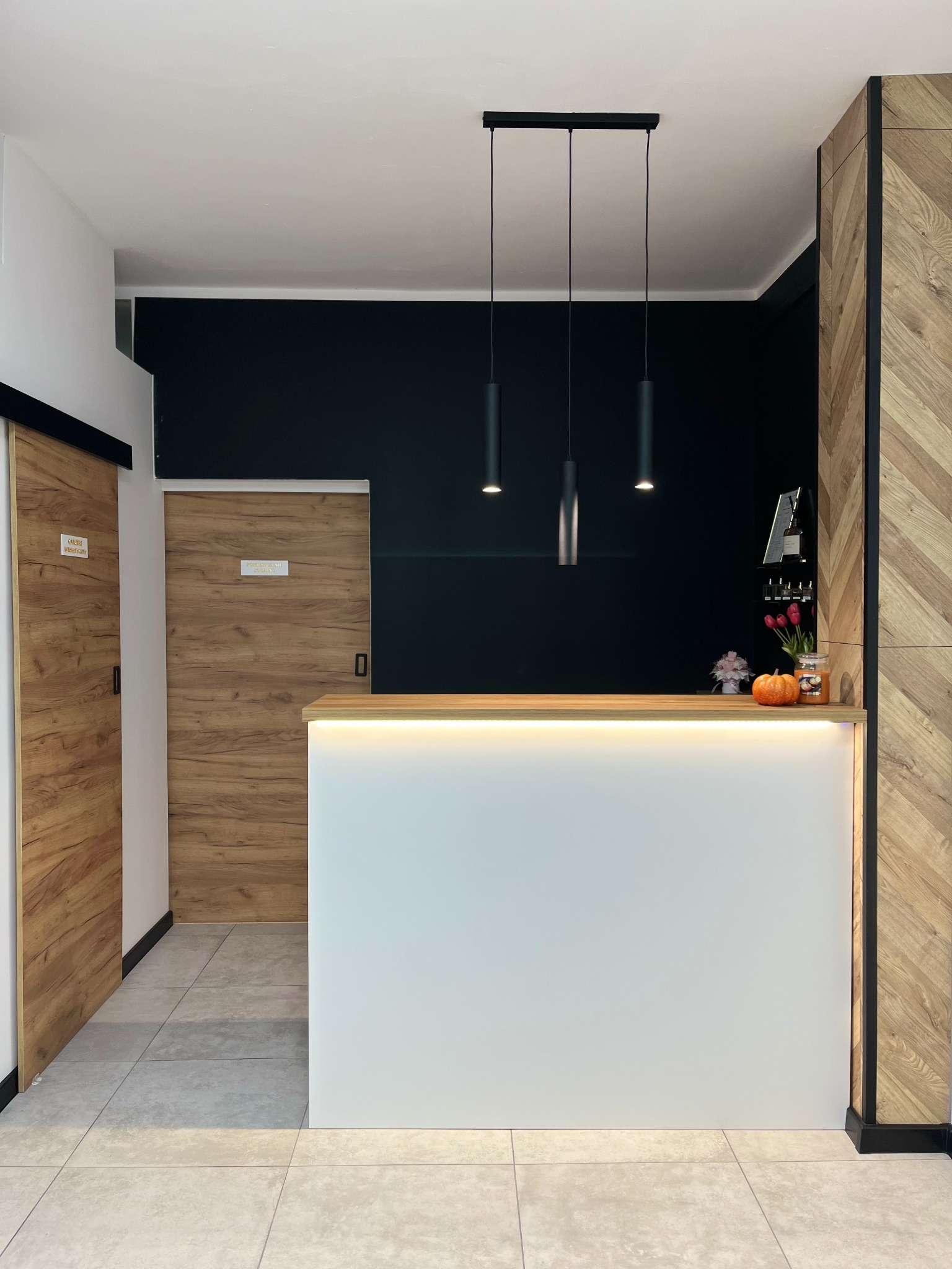 Recepcja z białym frontem podświetlonym ledami, drewnianym blatem, czarną ścianą w tle i trzema wiszącymi lampami. Po lewej stronie przesuwne drzwi z drewnianym wzorem, a po prawej ścianka...