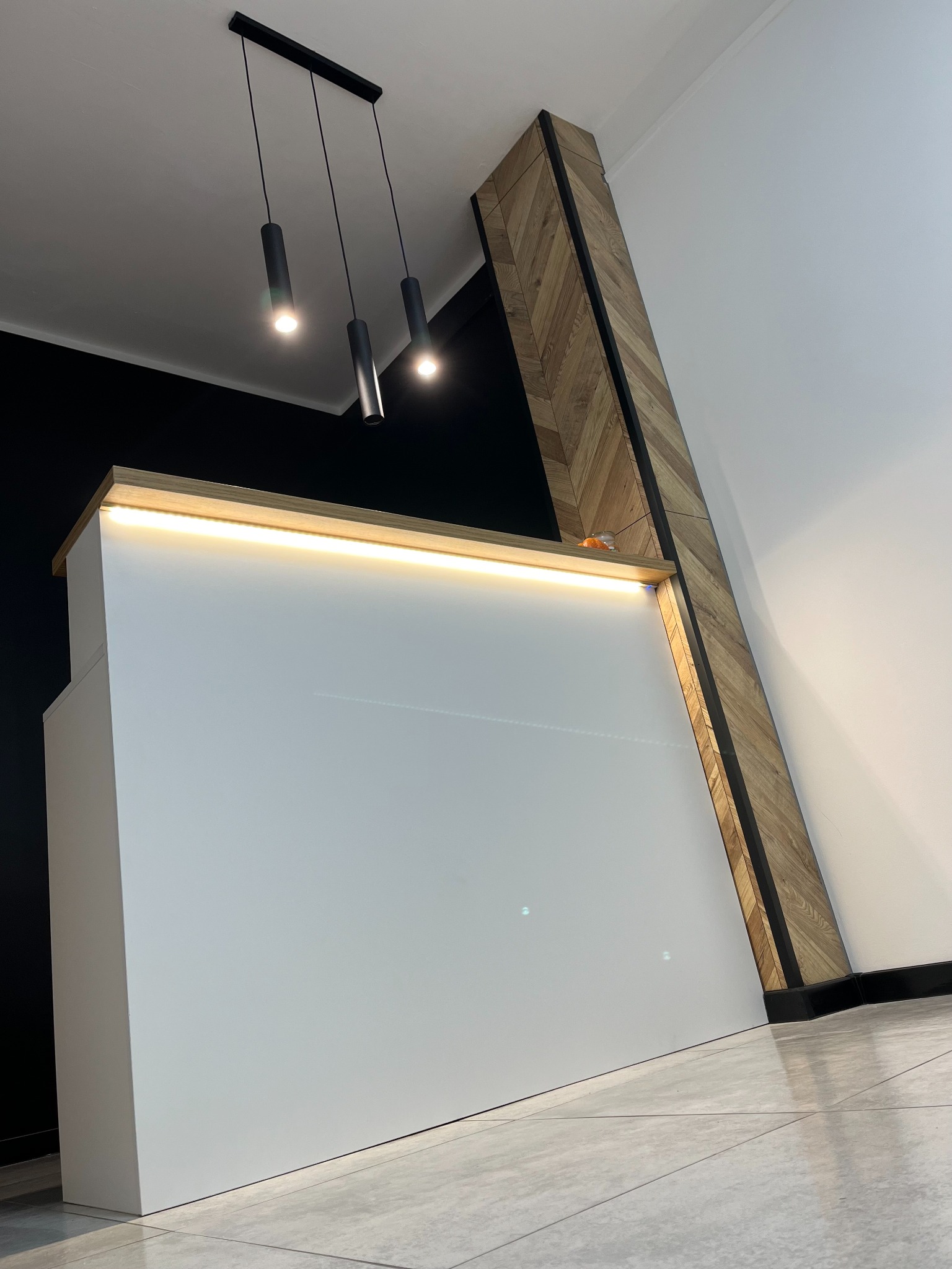 Biała, minimalistyczna zabudowa meblowa z podświetleniem LED i drewnianym akcentem w jodełkę, uzupełniona o czarne lampy sufitowe i szarą podłogę.