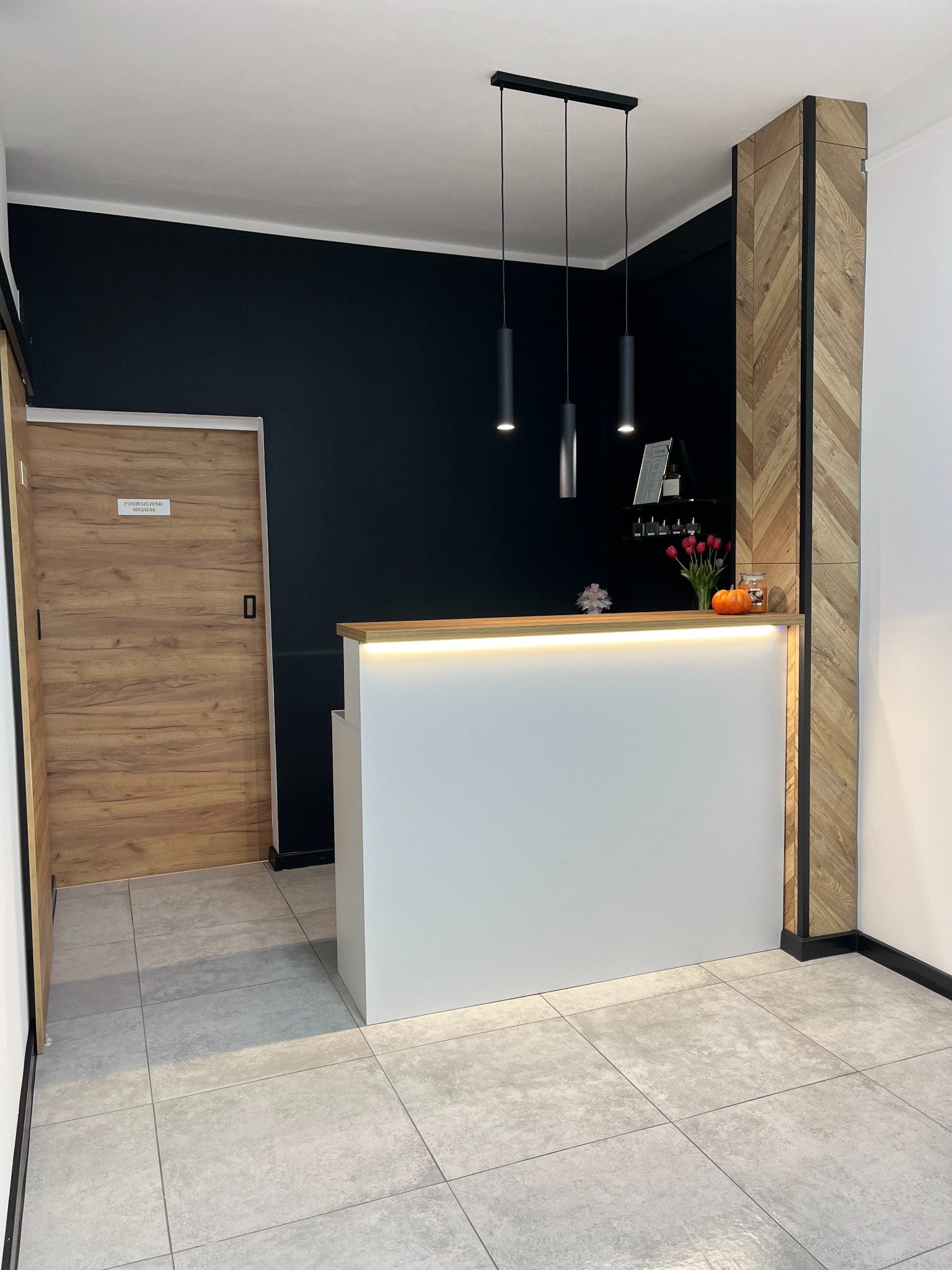 Recepcja z białym frontem, podświetlonym blatem z drewna, czarną ścianą w tle i elementem dekoracyjnym z drewna ułożonego w jodełkę.