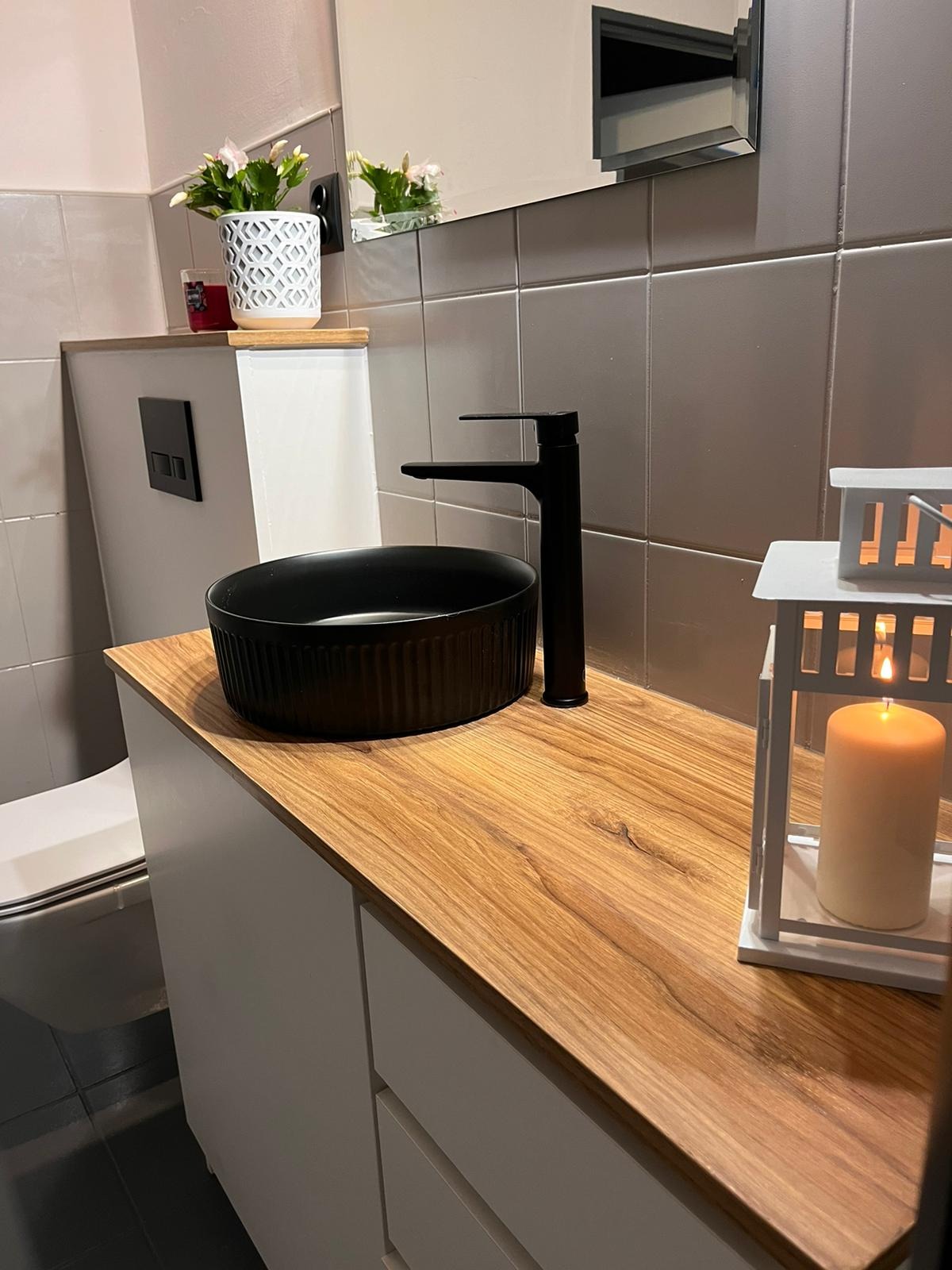 Łazienka z umywalką nablatową w kolorze czarnym, bateria w kolorze czarnym, blat drewnopodobny, biała szafka pod umywalką, toaleta podwieszana z czarnym przyciskiem, szare płytki na ścianie...