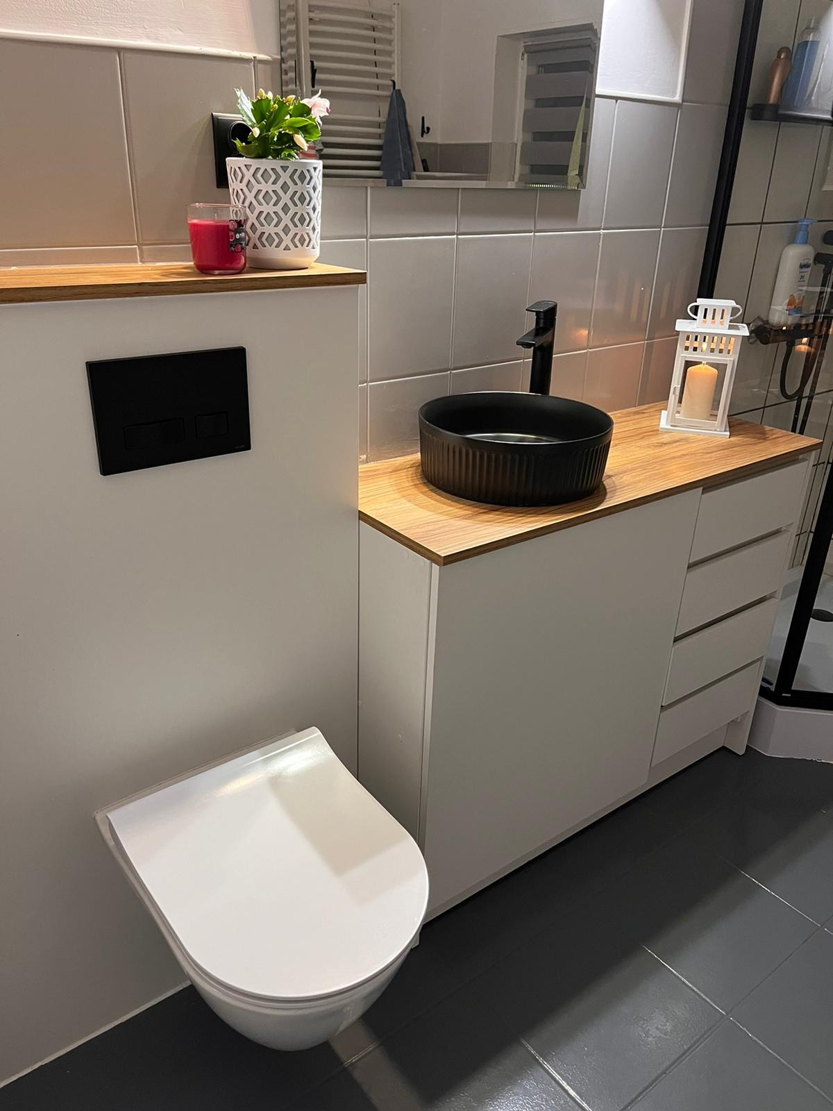 Nowoczesna łazienka z wiszącą toaletą, czarną umywalką nablatową i szafką z drewnianym blatem, uzupełniona dekoracjami: świecą, kwiatem i lampionem.