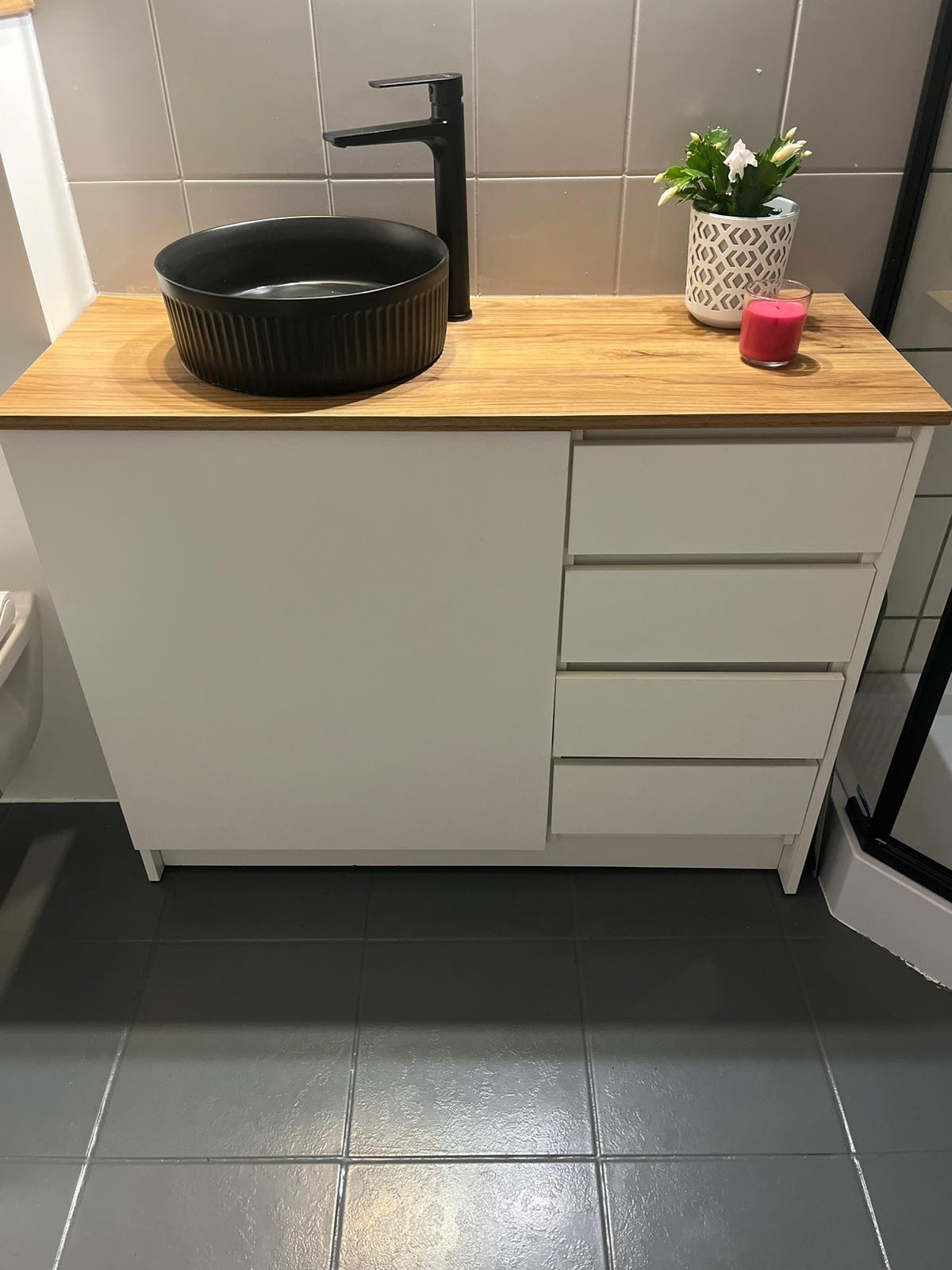 Łazienkowa szafka z białymi frontami i blatem z jasnego drewna, na którym stoi czarna umywalka nablatowa, czarna bateria, doniczka z kwiatami i różowa świeczka.