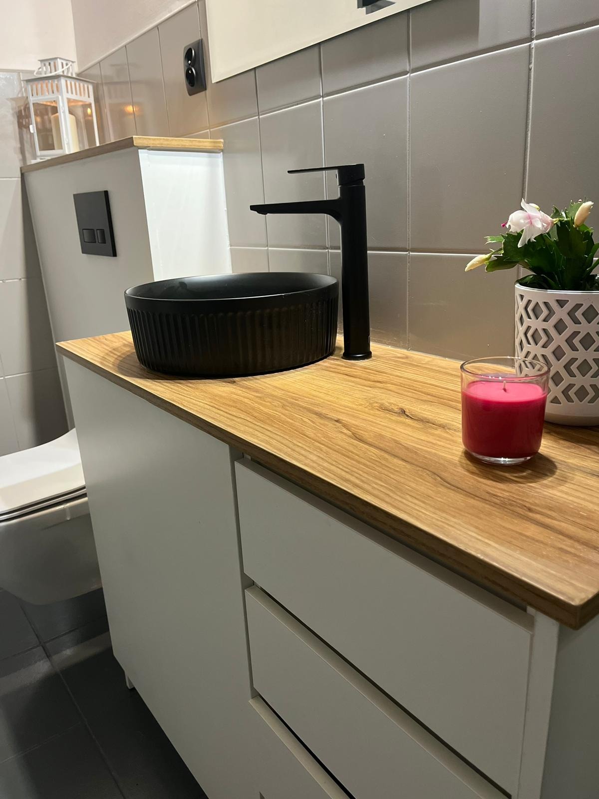 Czarna, okrągła umywalka nablatowa z czarną baterią na drewnianym blacie szafki łazienkowej z szufladami, obok świeczka i kwiat w ozdobnej doniczce.