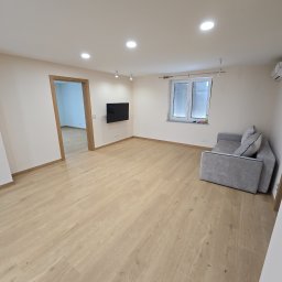 Jasne wnętrze po remoncie: drewniana podłoga, świeżo pomalowane ściany, okno z żaluzjami i szara sofa. Widoczne oświetlenie sufitowe i zamontowany telewizor.