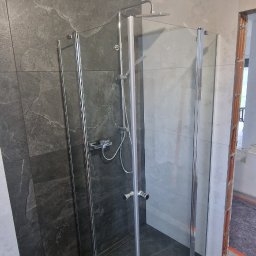 Nowoczesna kabina prysznicowa z chromowanymi profilami i deszczownicą, wykończona płytkami imitującymi kamień w odcieniach szarości. Widok z boku, wnętrze kabiny puste.