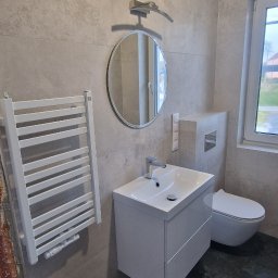 Damian kopczyński - Nowoczesna łazienka z umywalką na szafce, okrągłym lustrem i grzejnikiem drabinkowym. Szare płytki na ścianach i podłodze. Widoczne elementy armatury i toaleta.