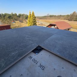 Szare płyty tarasowe ułożone na dachu z widocznym systemem montażowym i fragmentem membrany, w tle krajobraz z drzewami i budynkami w słoneczny dzień.