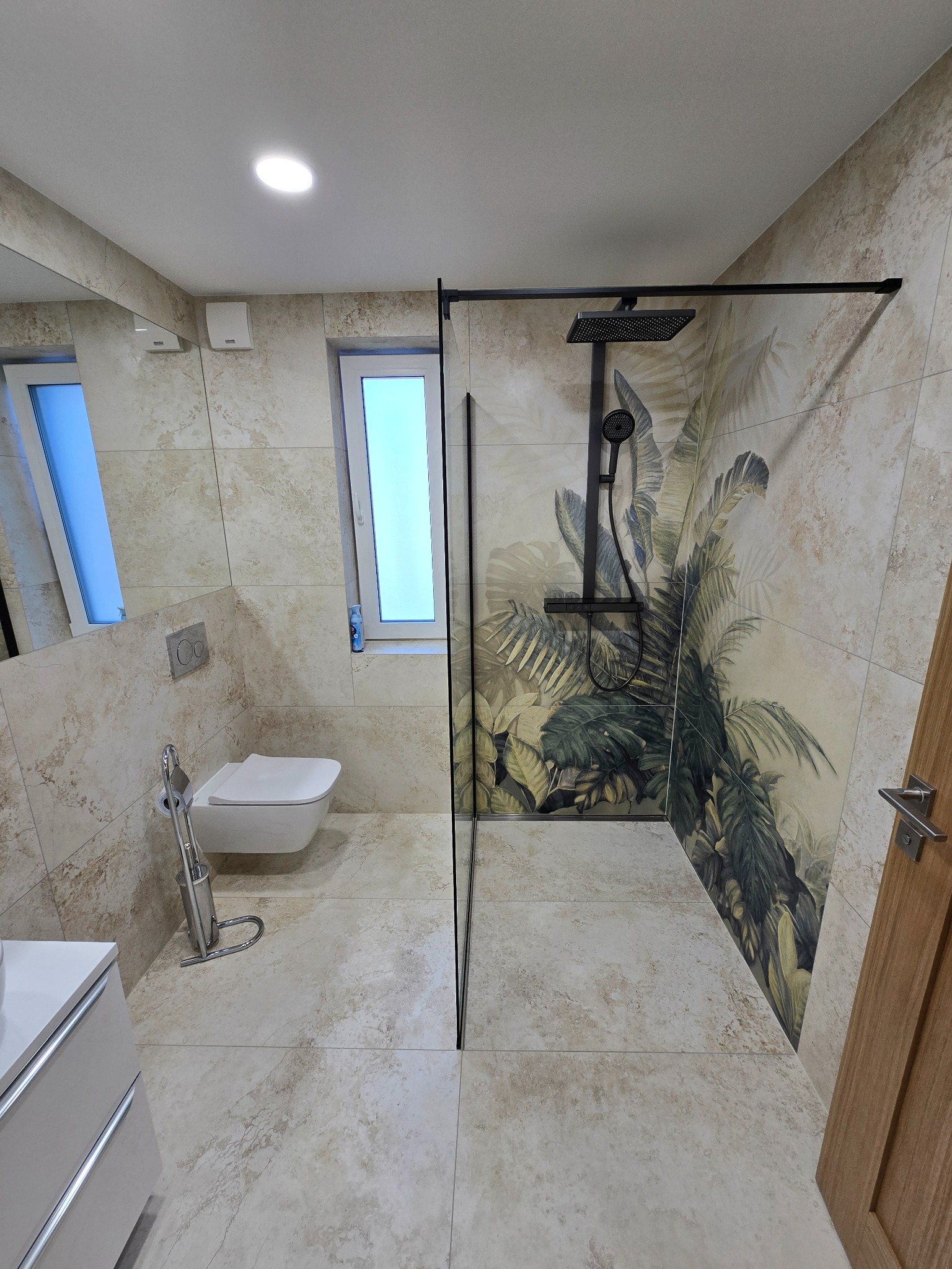 Nowoczesna łazienka z kabiną typu walk-in, beżowymi płytkami imitującymi kamień i akcentem tropikalnej tapety za prysznicem. Minimalistyczna toaleta pod oknem.