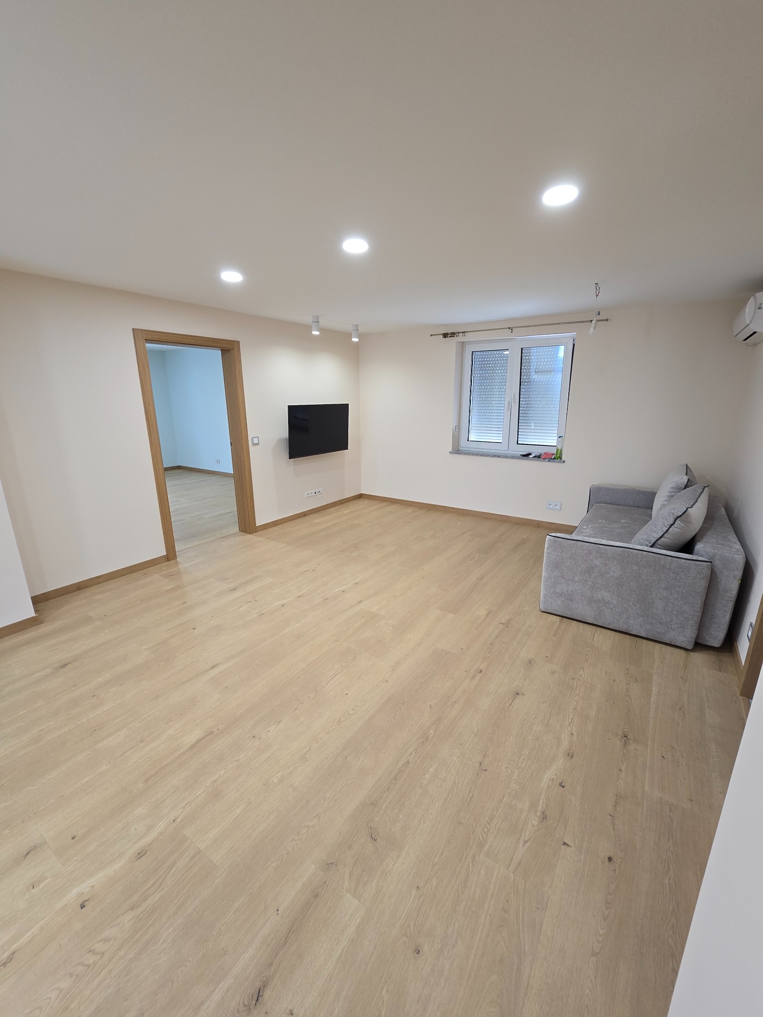 Jasne wnętrze po remoncie: drewniana podłoga, świeżo pomalowane ściany, okno z żaluzjami i szara sofa. Widoczne oświetlenie sufitowe i zamontowany telewizor.