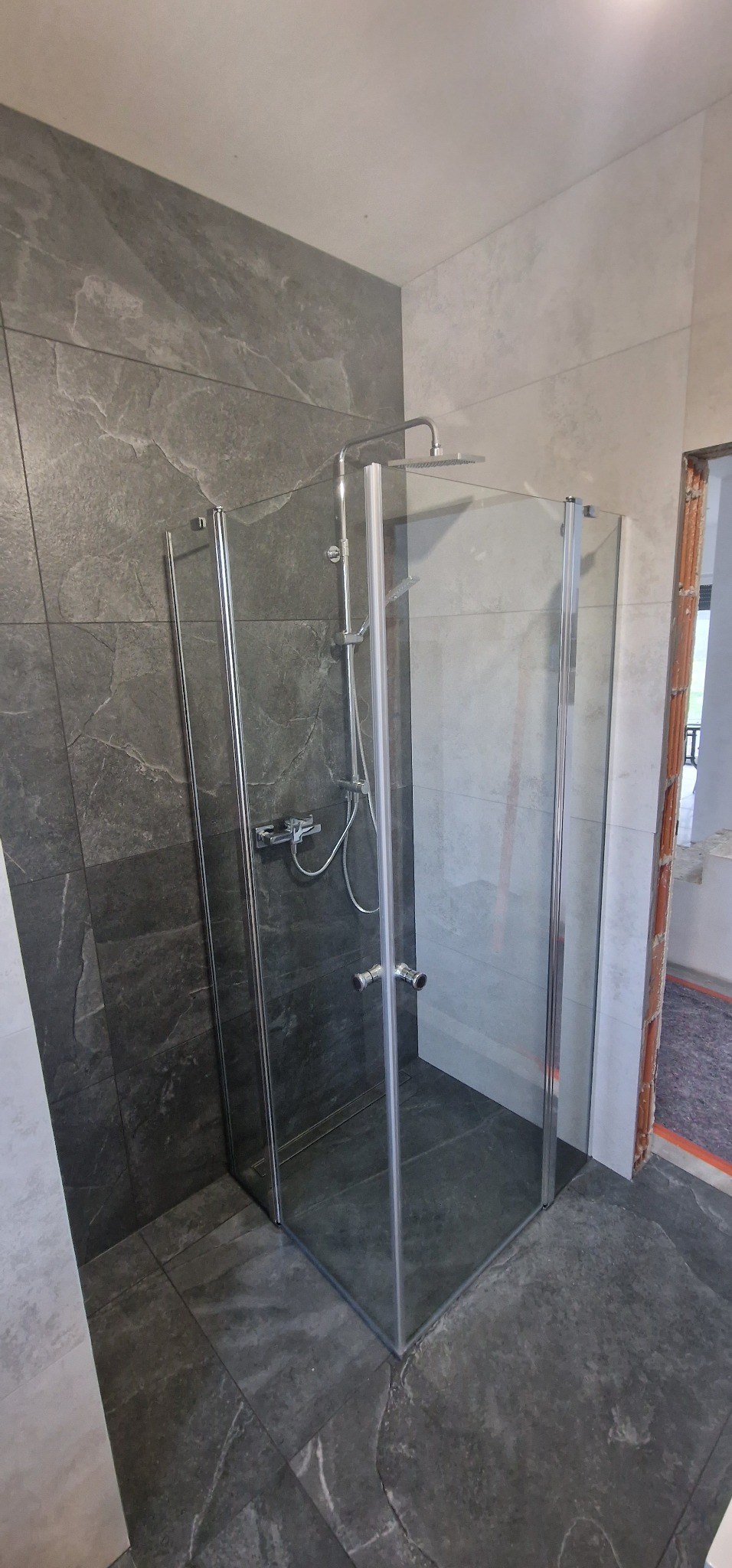 Nowoczesna kabina prysznicowa z chromowanymi profilami i deszczownicą, wykończona płytkami imitującymi kamień w odcieniach szarości. Widok z boku, wnętrze kabiny puste.