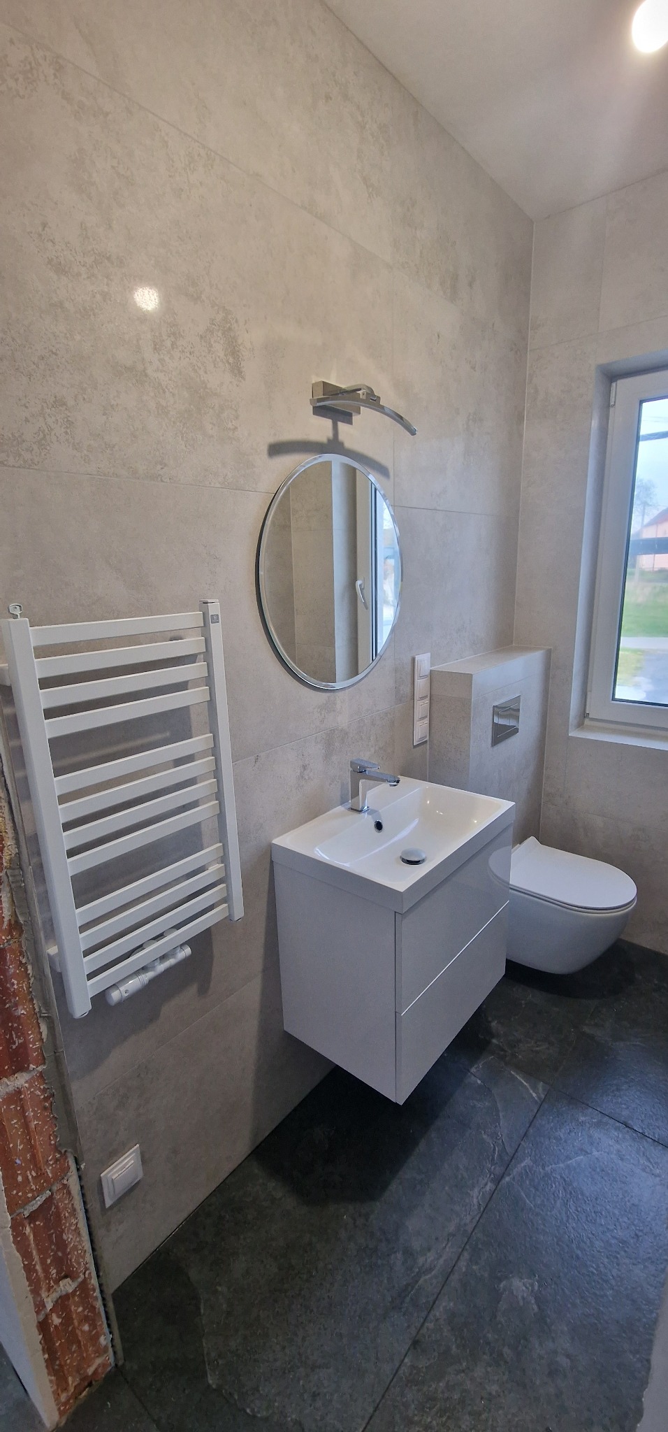 Nowoczesna łazienka z umywalką na szafce, okrągłym lustrem i grzejnikiem drabinkowym. Szare płytki na ścianach i podłodze. Widoczne elementy armatury i toaleta.
