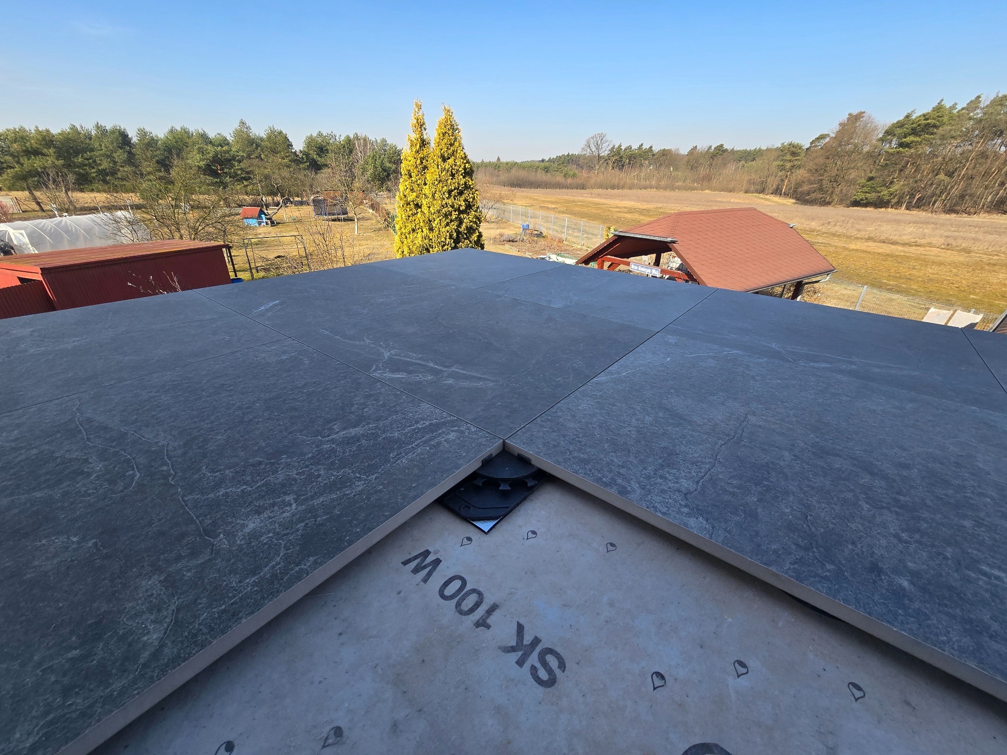 Szare płyty tarasowe ułożone na dachu z widocznym systemem montażowym i fragmentem membrany, w tle krajobraz z drzewami i budynkami w słoneczny dzień.