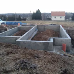 Zalane fundamenty domu jednorodzinnego, widok z góry na betonową konstrukcję, z instalacją i ziemią w środku. Na drugim planie zabudowa mieszkalna i krajobraz.