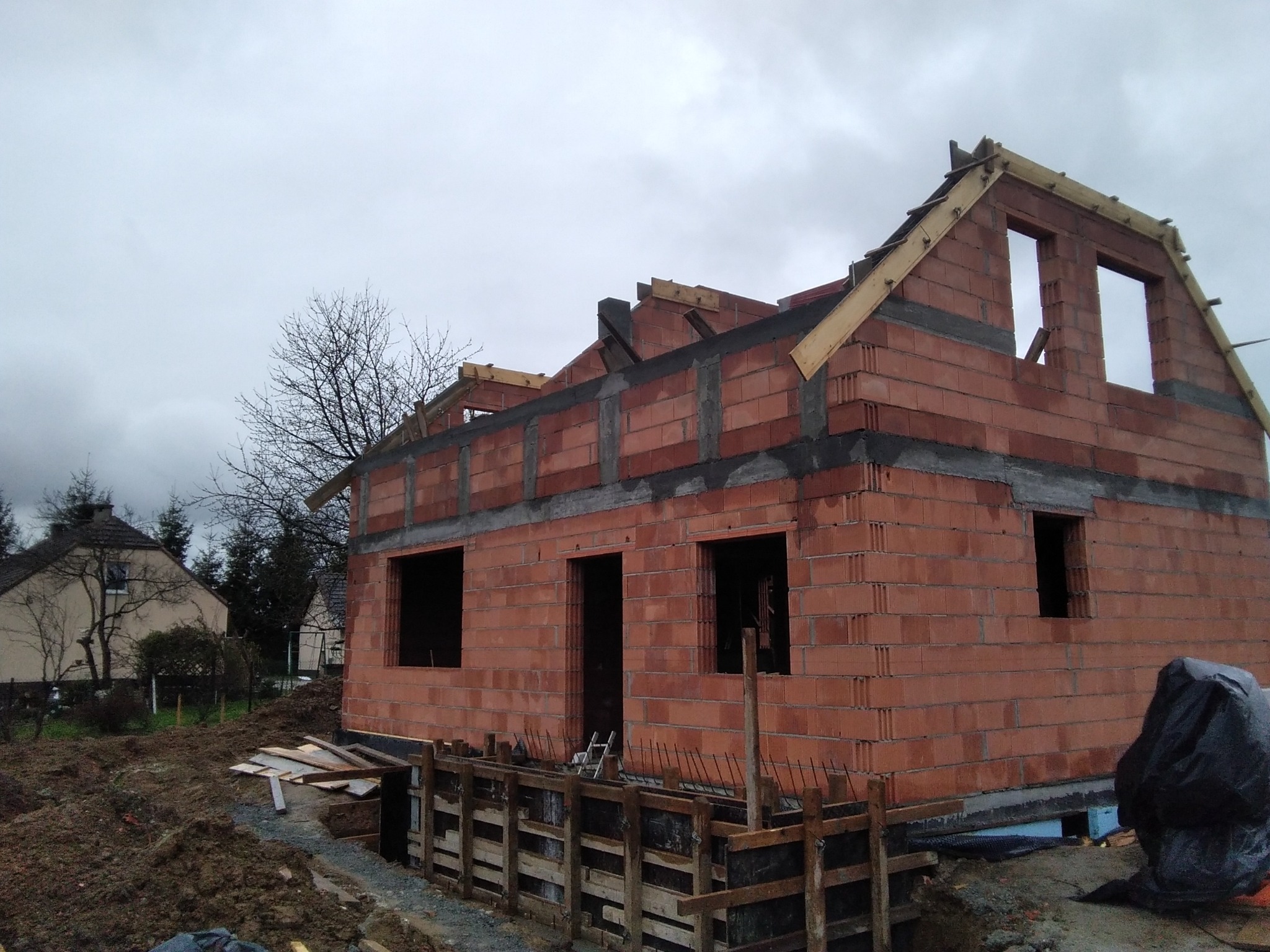 Surowy stan domu z czerwonej cegły z drewnianą konstrukcją dachu w trakcie budowy. Widoczne fundamenty i szare niebo w tle.