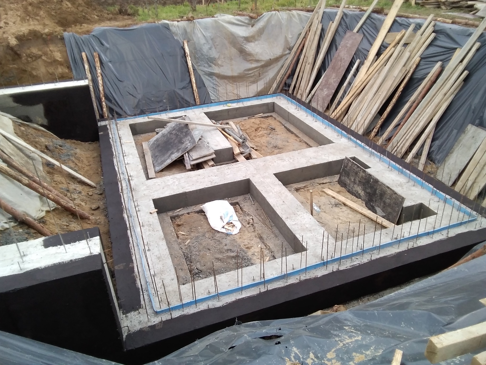 Betonowy fundament z hydroizolacją i wystającymi prętami zbrojeniowymi, otoczony wykopem i zabezpieczony folią. W tle drewniane stemple i deski.