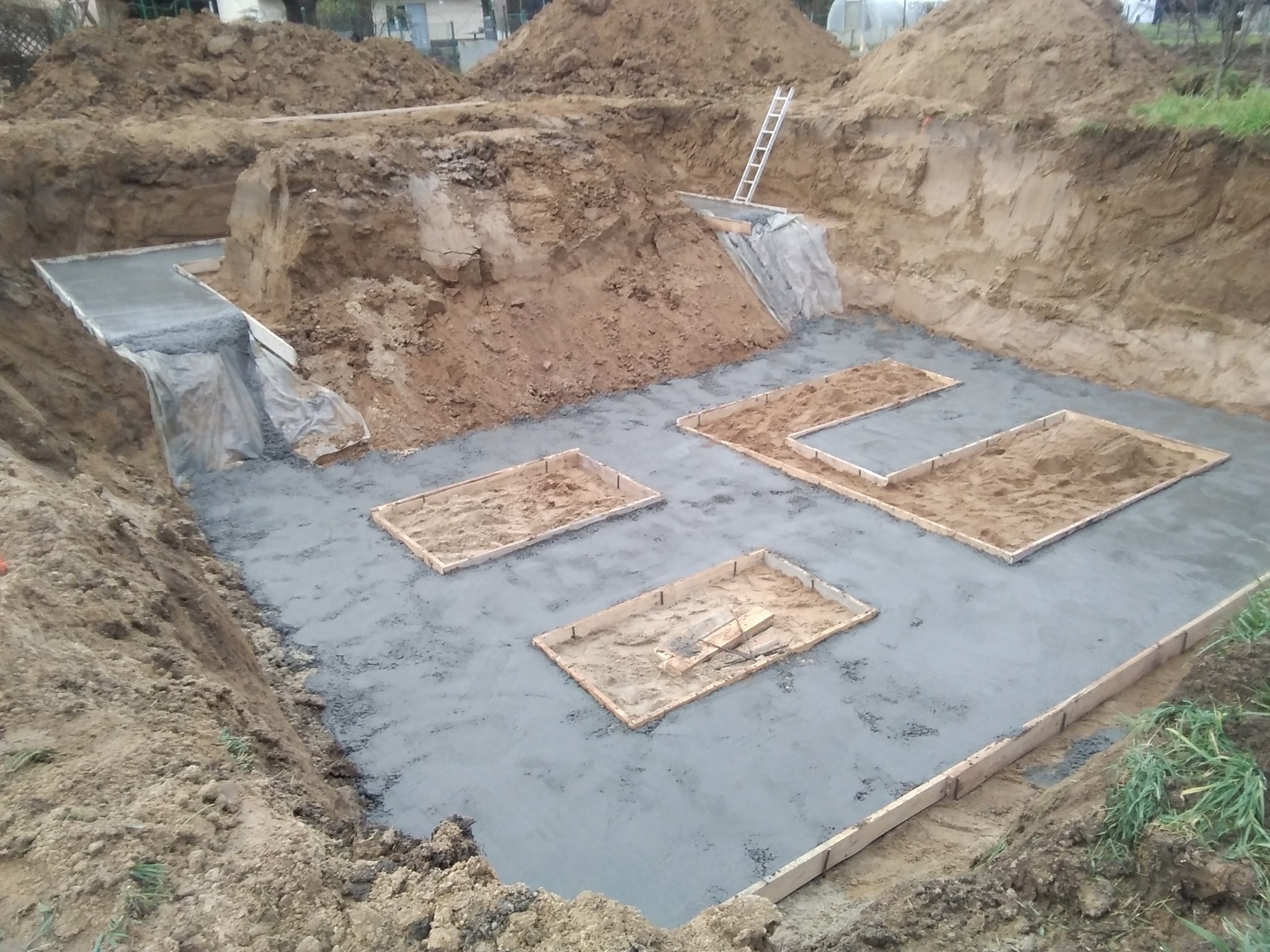 Zalany betonowy fundament z drewnianym szalunkiem i wykopaną ziemią w tle. Widoczny fragment drabiny oparty o skarpę ziemną.