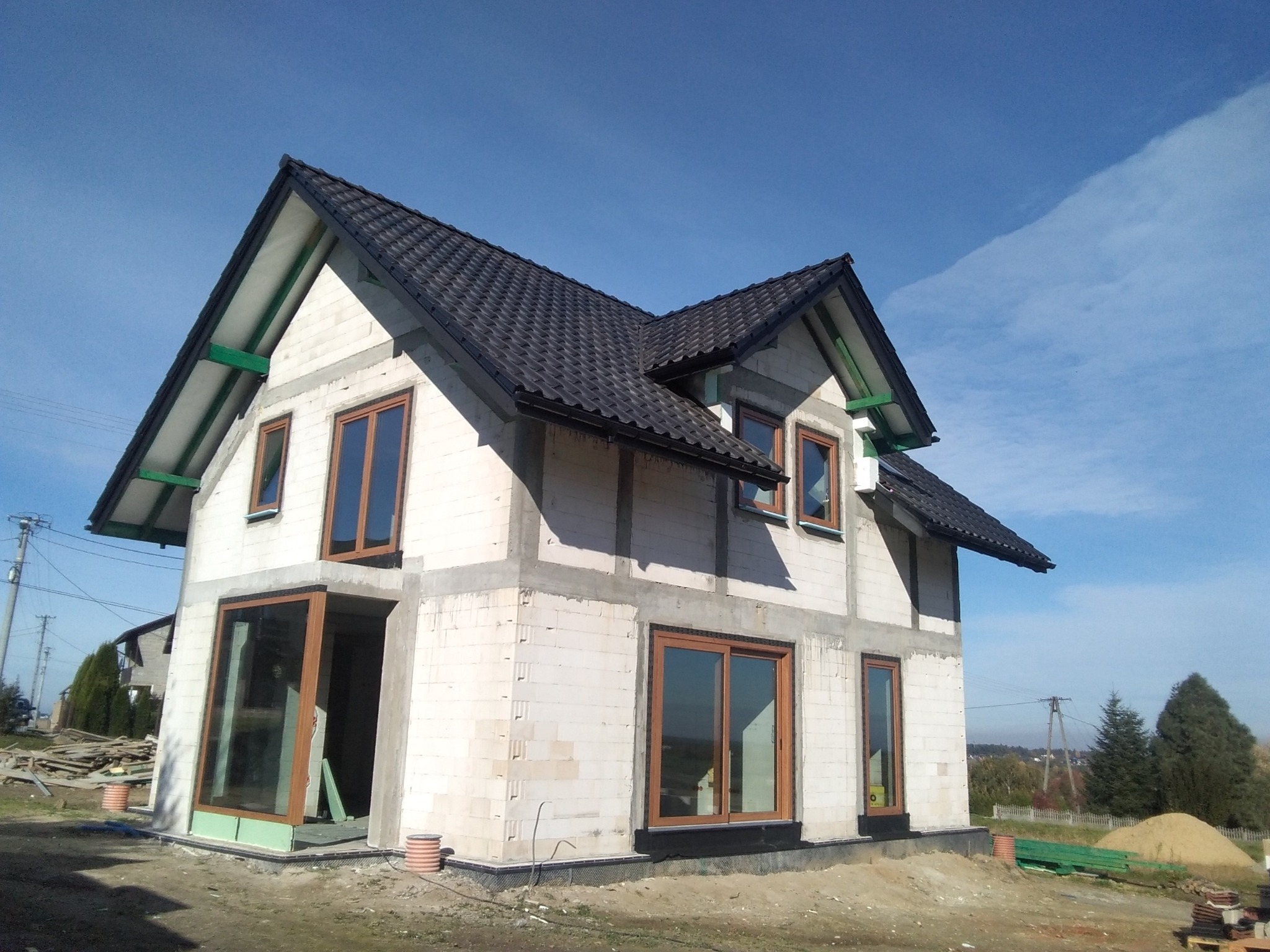 Surowy, dwukondygnacyjny dom z białego bloczka, z zamontowanymi oknami o drewnianych ramach i czarnym dachem, w trakcie budowy, na tle błękitnego nieba.