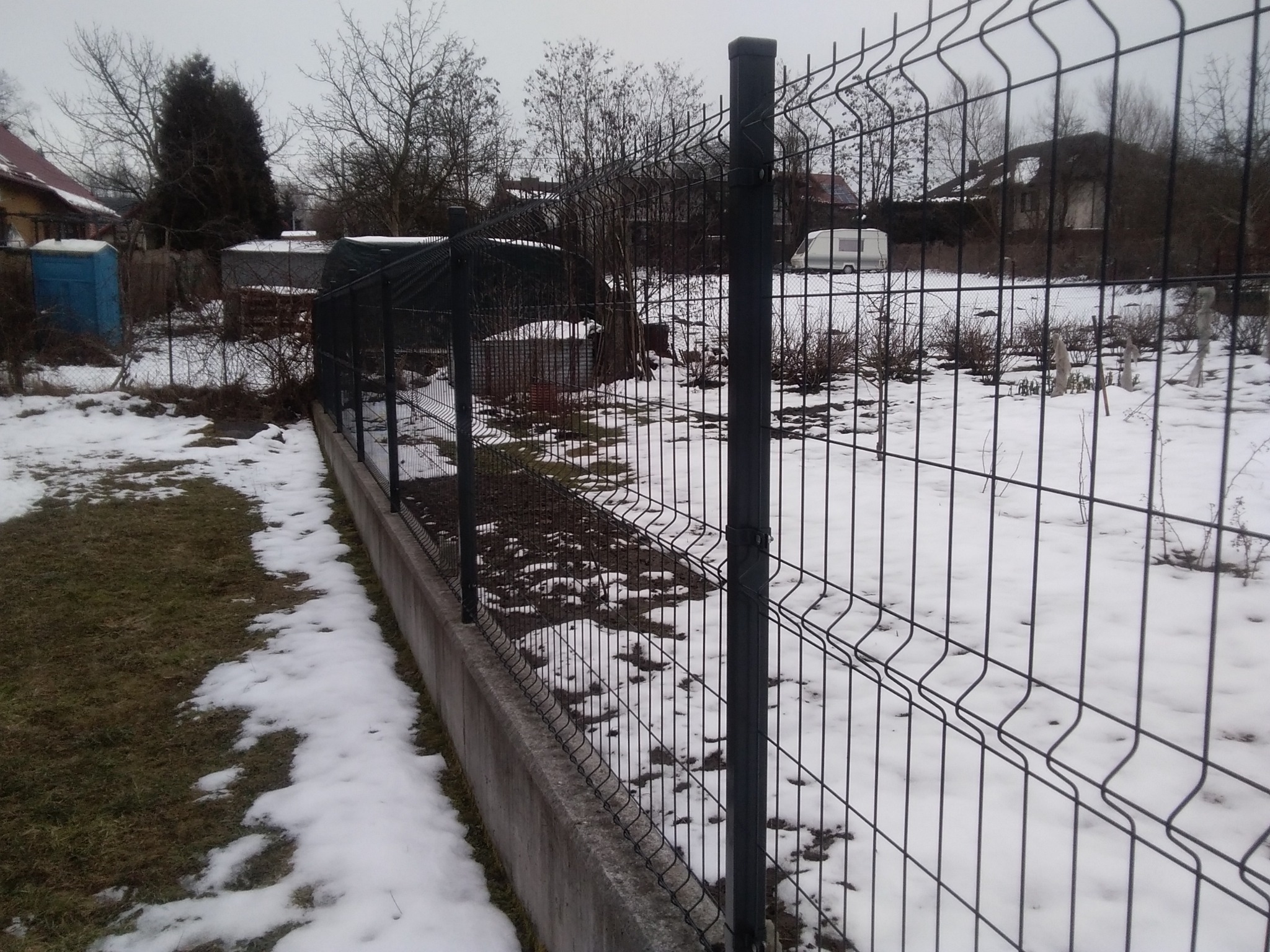 Ciemnoszare ogrodzenie panelowe na betonowej podmurówce, częściowo pokryte śniegiem. W tle zabudowania i drzewa bez liści, pochmurny dzień.