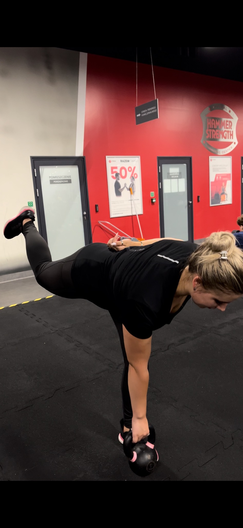 Kobieta w czarnym stroju sportowym wykonuje ćwiczenie równowagi z kettlebellem w siłowni Hammer Strength, unosząc jedną nogę do tyłu.