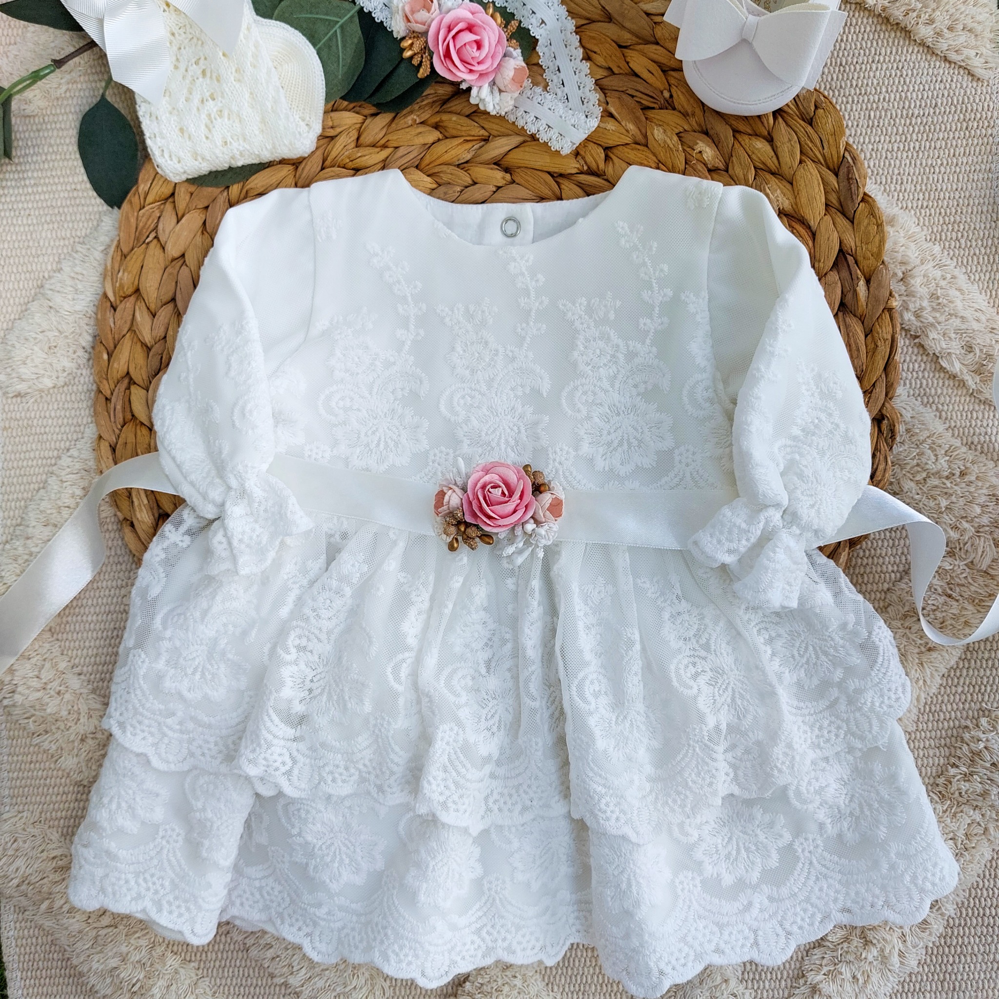 Biała, koronkowa sukienka niemowlęca w stylu Chanel z różową aplikacją kwiatową, na okrągłej, plecionej macie, z białymi bucikami, skarpetkami i opaską z kwiatami w tle.