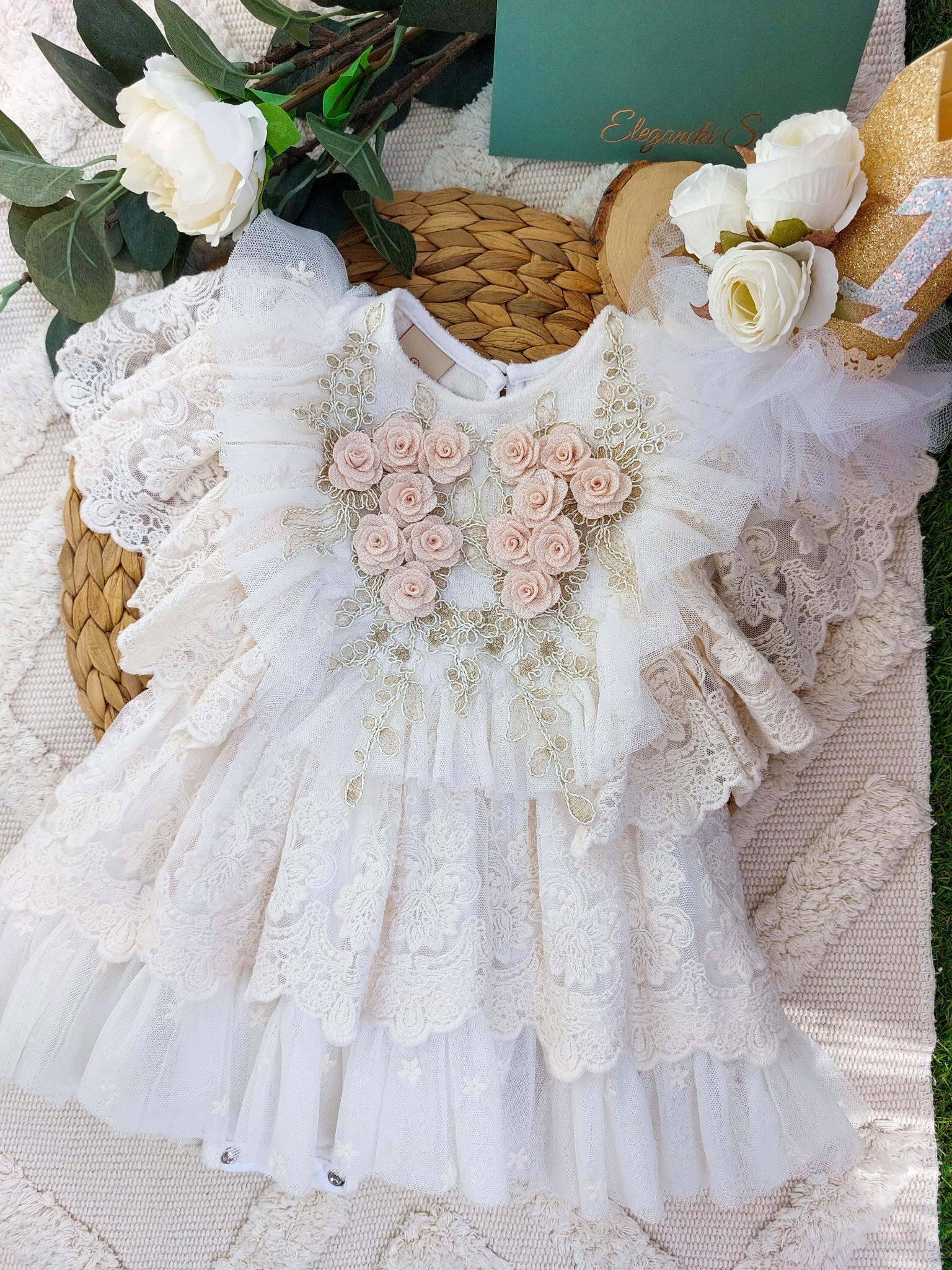 Elegancka, biała sukienka z koronkowymi falbanami i aplikacjami z różowych różyczek, leżąca na beżowym dywanie obok dekoracji z białych kwiatów i złotej korony z cyfrą jeden.