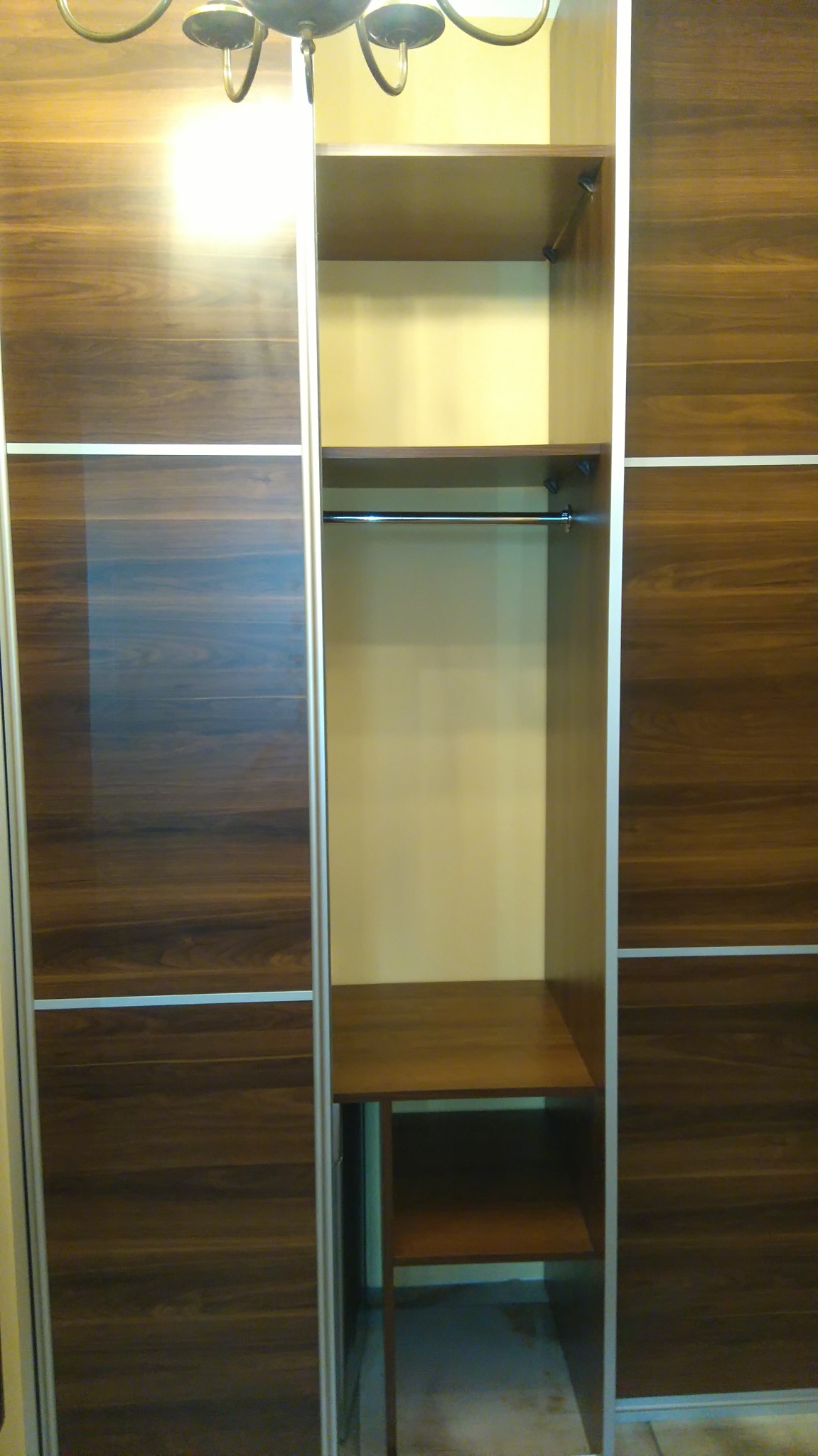 Szafa przesuwna z drewnopodobnymi frontami, widoczna pusta przestrzeń z półkami i drążkiem na ubrania, oświetlenie sufitowe w górnej części kadru.