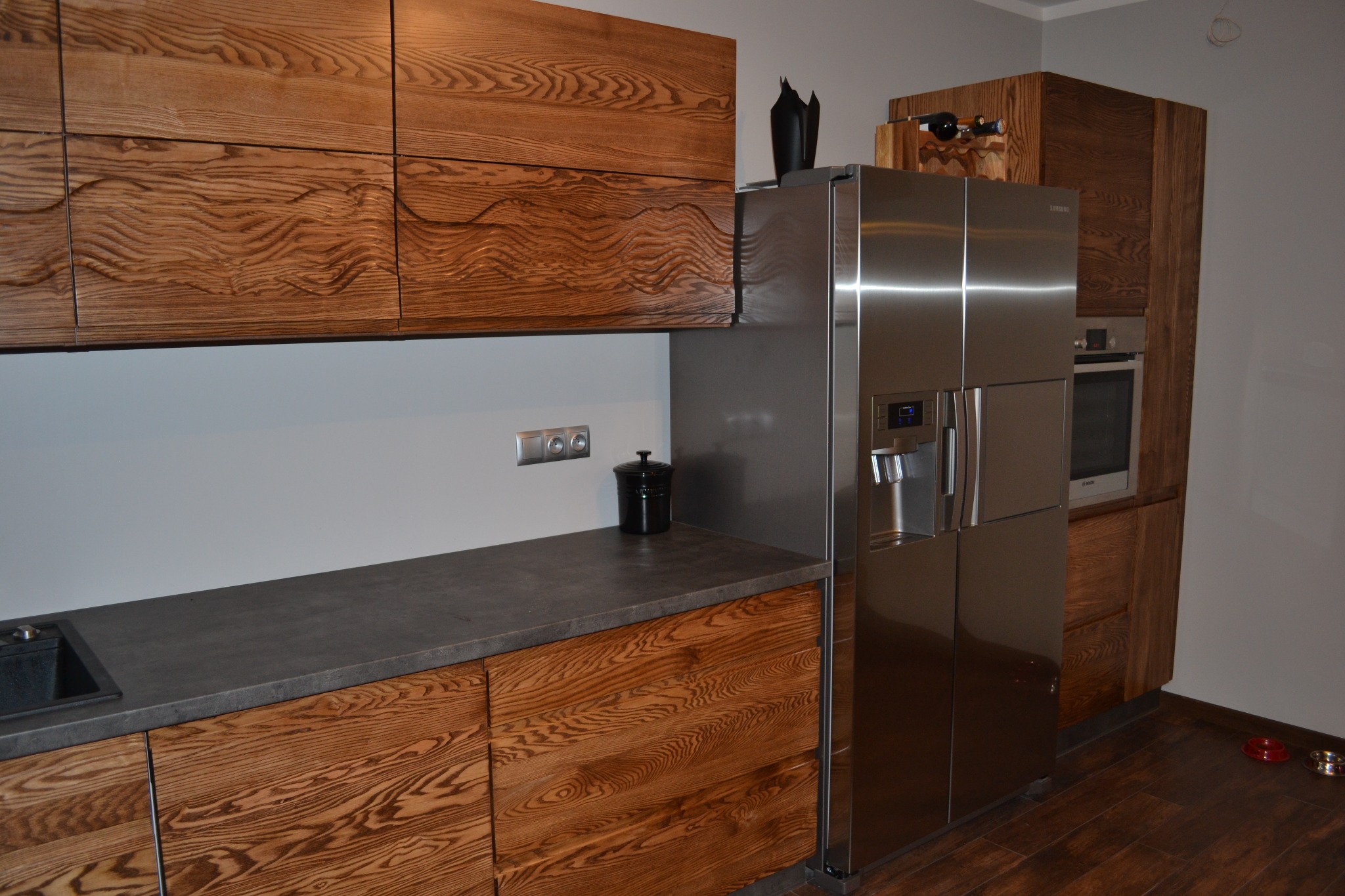 Nowoczesna kuchnia z drewnianymi frontami o wyraźnym usłojeniu, szarym blatem i dużą, dwudrzwiową lodówką ze stali nierdzewnej. Na blacie czarny pojemnik, a nad lodówką drewniany stojak na wino.