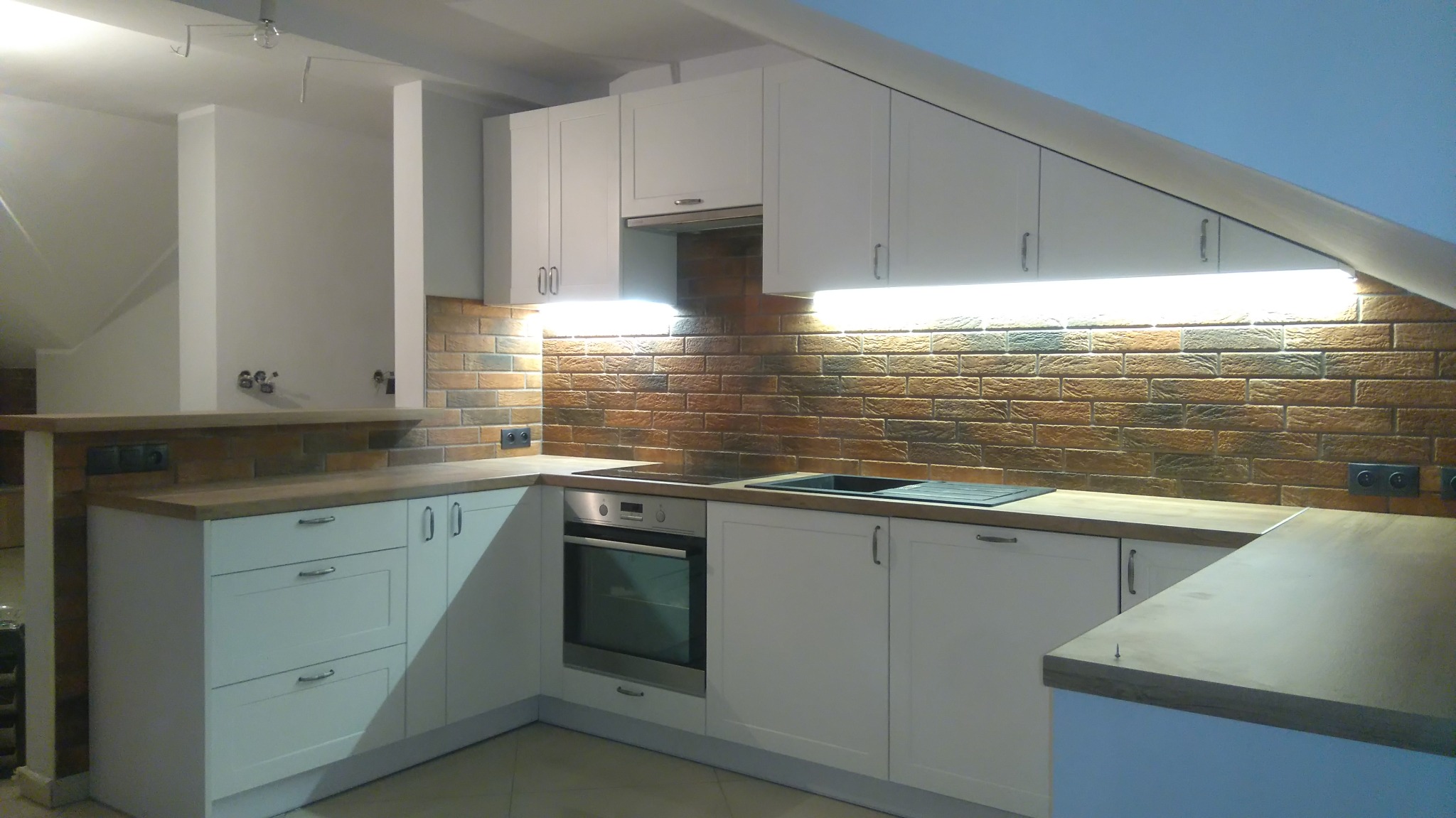 Biała kuchnia w kształcie litery L w pomieszczeniu ze skosem sufitowym, z drewnianym blatem i ceglaną ścianą, oświetlona listwą LED pod szafkami.