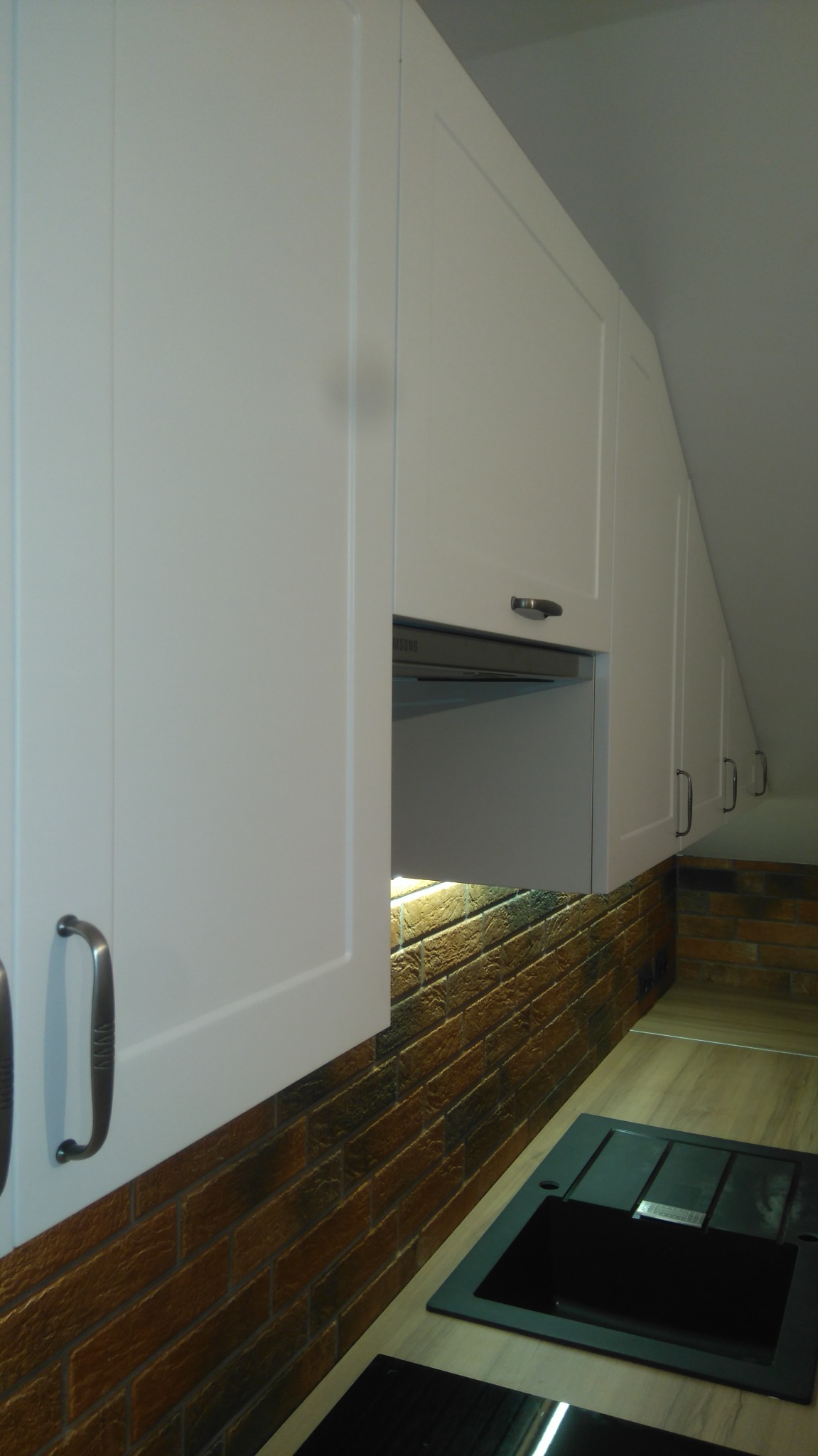 Białe szafki kuchenne pod skosem sufitu, z metalowymi uchwytami, oświetleniem podszafkowym i ceglaną ścianą nad blatem z czarnym zlewem.