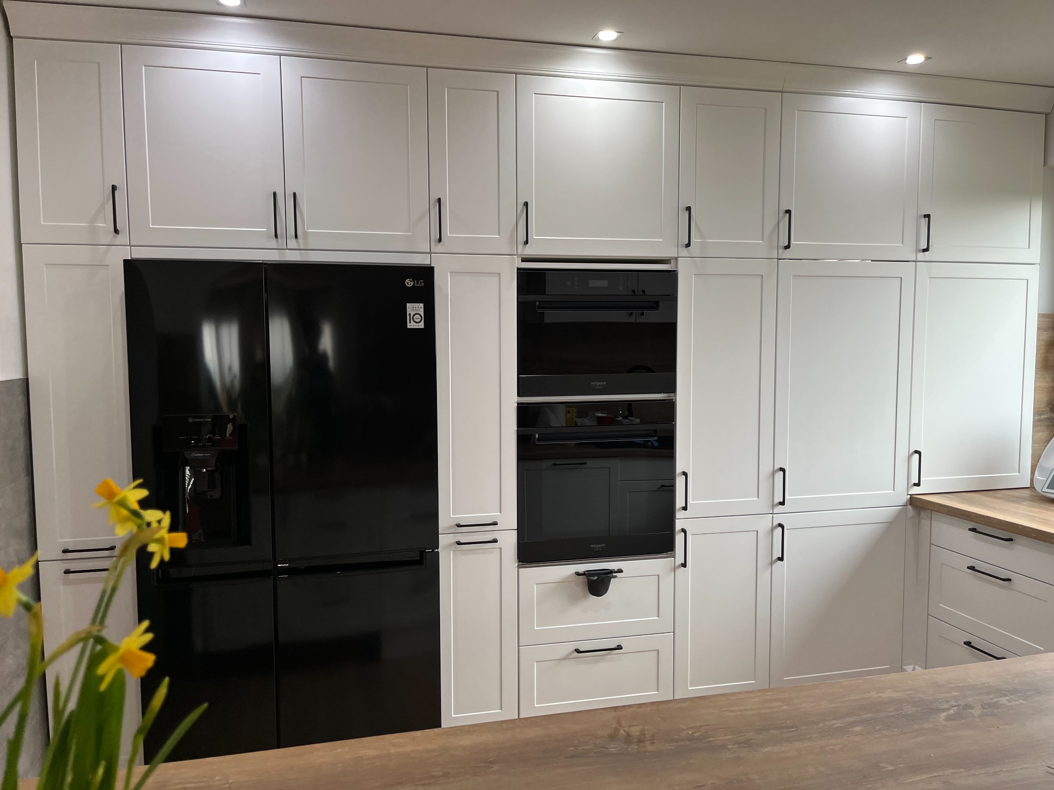 Biała zabudowa kuchenna z czarnym sprzętem AGD, minimalistyczne uchwyty, drewniany blat w tle, bukiet żółtych narcyzów na pierwszym planie.